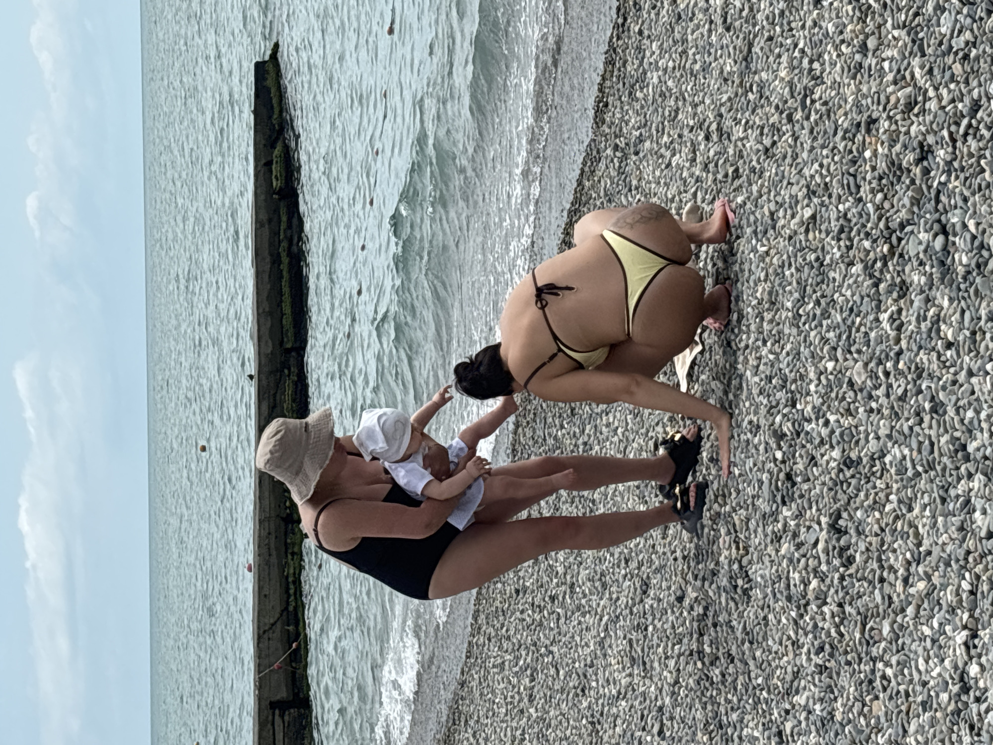 The image shows a pebble beach in Sochi, Russia, with two women and a baby near the water's edge. The woman on the left is wearing a black swimsuit and a light-colored bucket hat, holding a baby dressed in white. The baby is facing away from the camera, towards the woman on the right. The woman on the right is wearing a yellow bikini and is crouched down, facing the baby. The water is a light blue-grey and appears to be moderately wavy. In the background, a concrete pier extends into the water, and buoys are visible further out. The sky is overcast with some white clouds. The ground is covered with small, grey pebbles.