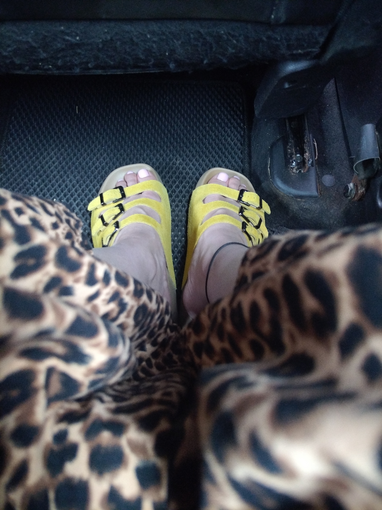 In Zhodzina, Belarus, a close-up, high-angle shot shows a pair of feet wearing yellow slide sandals with black buckles. The feet are resting on a textured black floor mat inside a vehicle. The person is wearing a garment with a leopard print pattern that drapes over their legs. The background is dark and appears to be the interior of a car, with a glimpse of a dark grey car seat and some car components on the right side.