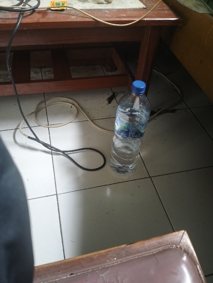 The image shows a room in a house in Ciparay, Indonesia, with a wooden table, a blue plastic water bottle, and a black electrical cord. The water bottle is sitting on the floor in front of the table, which has a shelf underneath. The electrical cord is coiled up on the floor and appears to be plugged into the table. The floor is tiled and has some dirt marks. The walls and part of a bed are visible in the background. The scene is quiet and appears to be undisturbed.