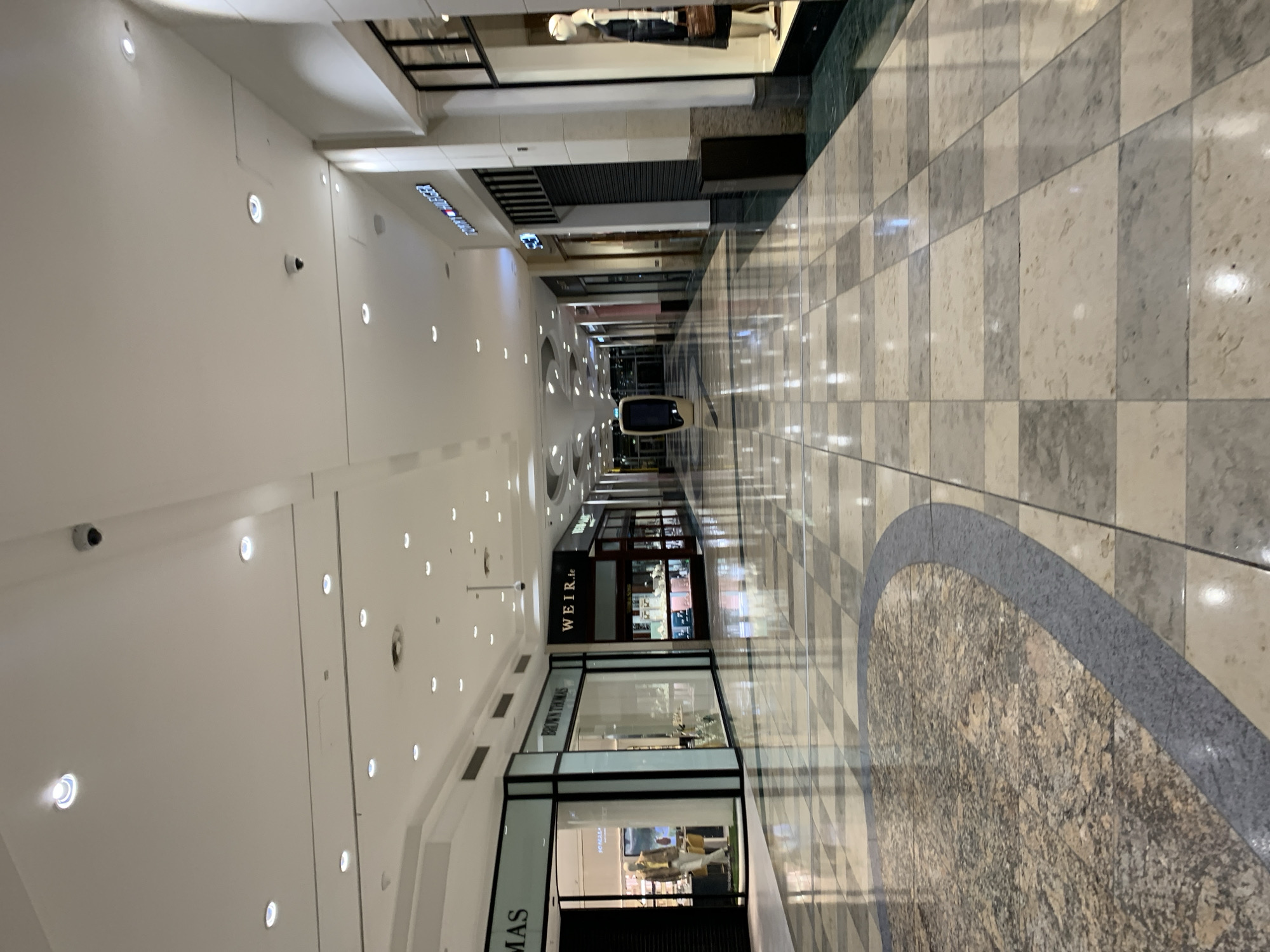 This is an indoor view of a shopping mall hallway. The hallway has a white ceiling with evenly spaced recessed lights. Security cameras are also visible along the ceiling. Shopfronts line both sides of the hallway, including "WEIR.ie" and "BROWN THOMAS." The floor is tiled in a checkered pattern of alternating light and dark squares, with a decorative circular pattern in the center. At the end of the hallway, a single vehicle is visible. Based on the visible retailers, this photo appears to be taken inside Dundrum Town Centre, located in Dundrum, Dublin 16, Ireland. No people are visible in the photo.