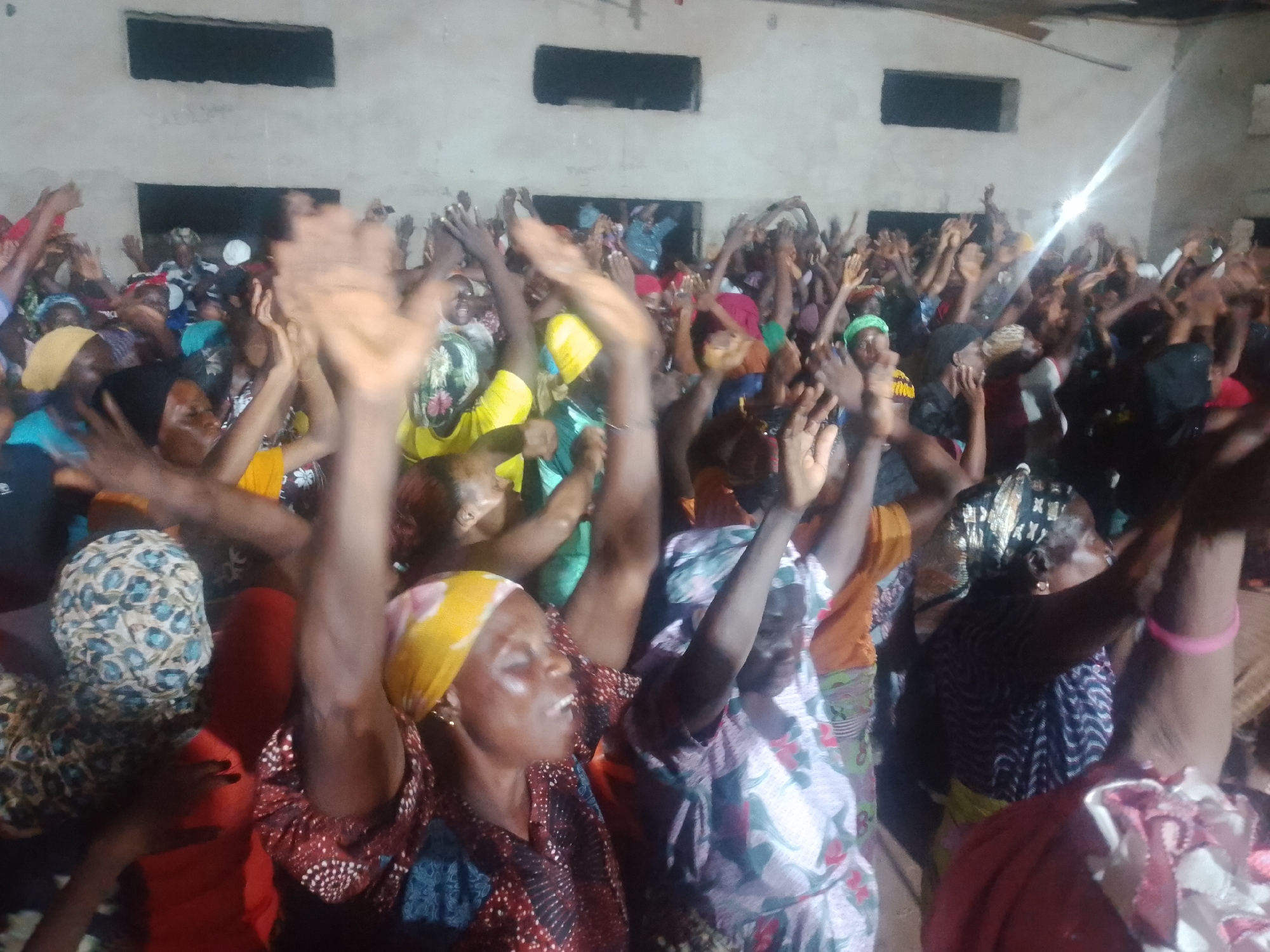 The image depicts a dimly lit interior scene with a large group of people, predominantly women, with their arms raised in the air. The setting appears to be a communal gathering or event, possibly a religious service or celebration, given the raised hands and expressions of emotion on some faces. The walls are rough and unpainted, with two rectangular openings that might be windows or ventilation shafts. The lighting is uneven, with a bright beam of light emanating from the upper right, suggesting a flash or a strong artificial light source. The people are dressed in vibrant, patterned clothing, including headscarves and dresses. Some individuals are clapping, while others have their eyes closed or are looking upwards. The overall impression is one of energetic participation and communal engagement within a simple, somewhat rustic environment.