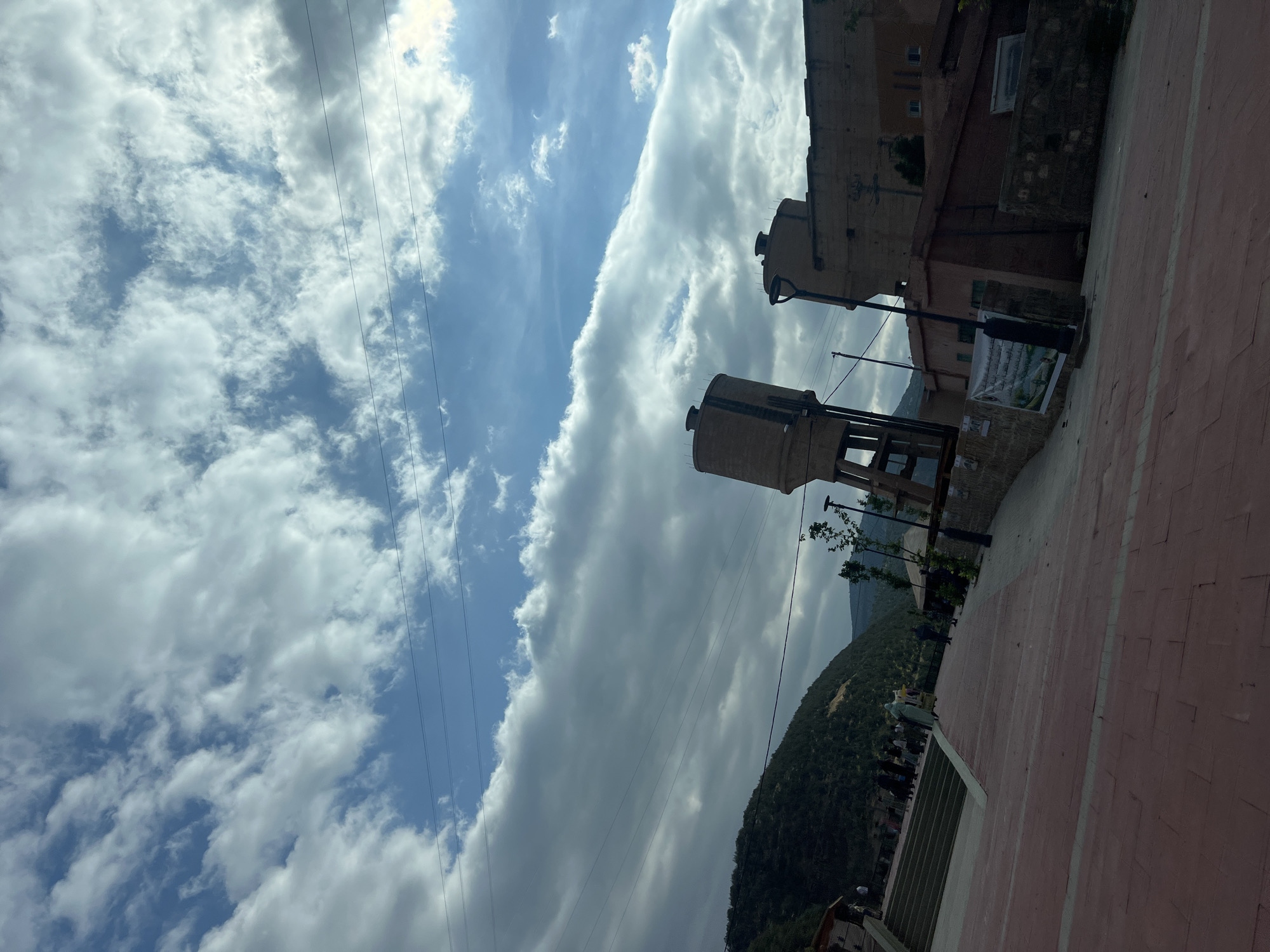 The image depicts a scene in Bin El Ouidane, Morocco. Two cylindrical water towers are visible on a metal support structure, situated next to a building of indeterminate function. A paved area, possibly a walkway, extends into the foreground. In the background, a hill covered with vegetation is present. The sky is filled with a mix of blue sky and clouds. Overhead power lines cut across the sky.