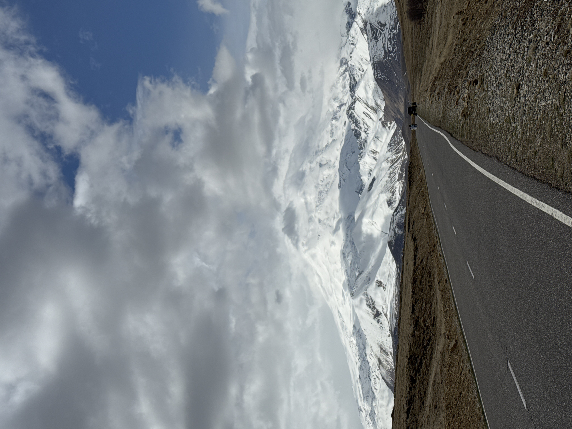 A long, straight asphalt road with a solid white line running down the right side dominates the scene. The road is bordered by a rocky, dirt embankment on the right side. A person is visible in the distance, standing on the road. To the left of the road are snow-covered mountains that rise dramatically. The sky above the mountains is filled with white and grey clouds. A small patch of blue sky is visible in the upper left corner. The photo gives the impression of a mountain pass or scenic route in Unknown, Russia.