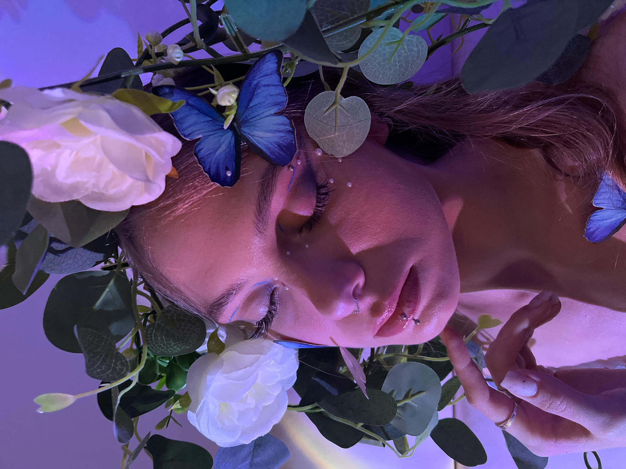 A close-up, tilted portrait shows a person with a floral and butterfly adorned headpiece, eyes closed. The person has pale skin, light brown hair, and light blue makeup accents their eyes. Small, white bead-like embellishments are scattered on their forehead. A blue butterfly is positioned near their left eye. They have piercings in their nose and lip. Their hand, wearing a ring and showing a painted nail, is partially visible near their face. The surrounding foliage includes white roses and eucalyptus leaves. The background is a gradient of light purple. No discernible details indicate a location in the city of Фоки, Russia.