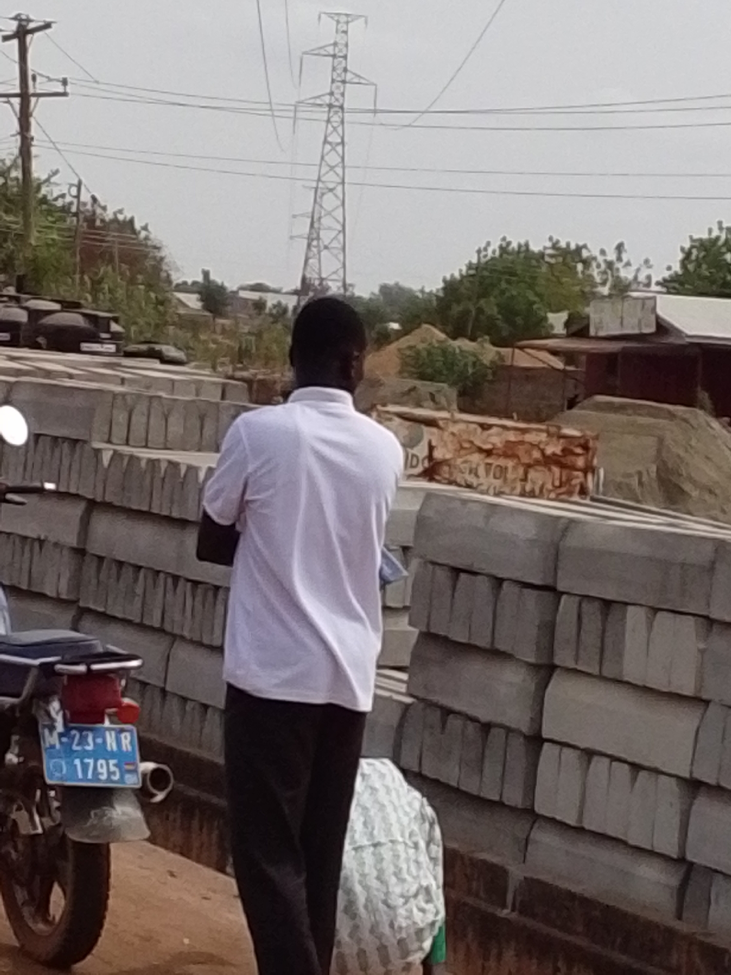 A man in a white short-sleeve collared shirt and dark pants stands near a stacked pile of gray, concrete blocks. The man is facing away from the camera and towards the blocks. Next to the blocks is a motorcycle. The license plate reads "M-23-NR 1795 GH." Behind the blocks are piles of sand and some brick debris. In the background, an electrical tower is visible against a gray sky. This scene is located in the city of Tamale, Ghana.