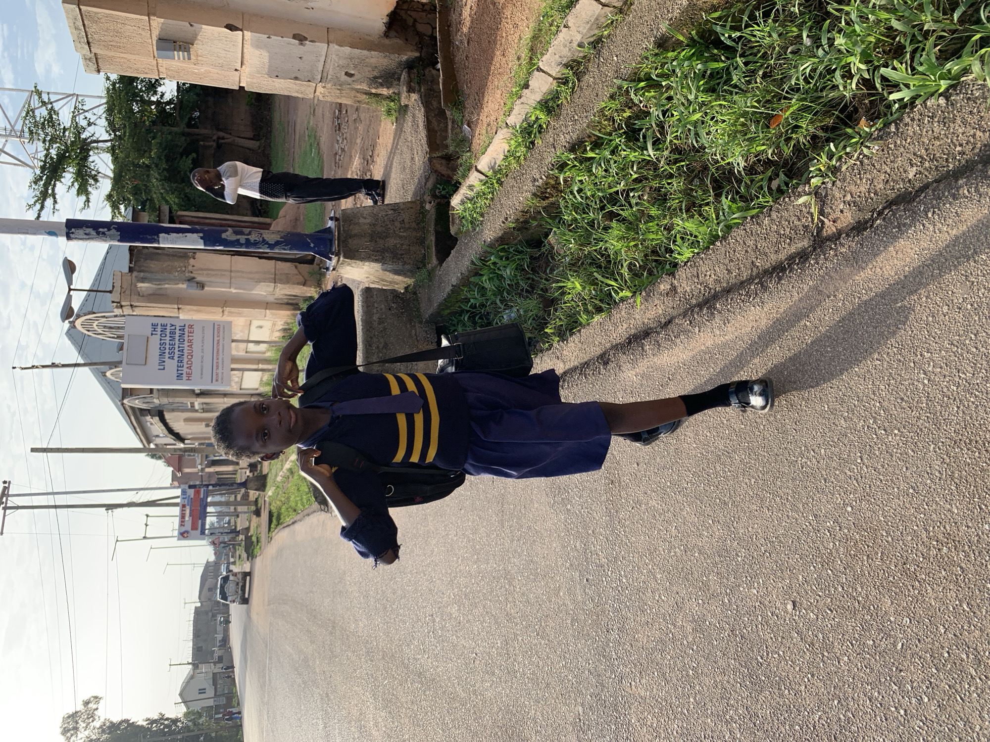 A young Black girl, wearing a school uniform with a navy blue skirt and a navy blue top with yellow stripes on the sleeves and chest, walks on the side of a paved road in Jos, Nigeria. She has a black backpack slung over her shoulders and a black bag in her right hand. Her hair is short and dark, and she is looking forward. The road is bordered by a drainage ditch filled with green grass and some weeds. In the background, a man in a white and black shirt and black pants stands with his hand on his head. There are buildings and utility poles visible along the road, suggesting an urban or semi-urban environment. A sign on a building reads "THE LIVINGSTONE ASSEMBLY INTERNATIONAL HEADQUARTER." The lighting indicates it is daytime, with the sun casting shadows.