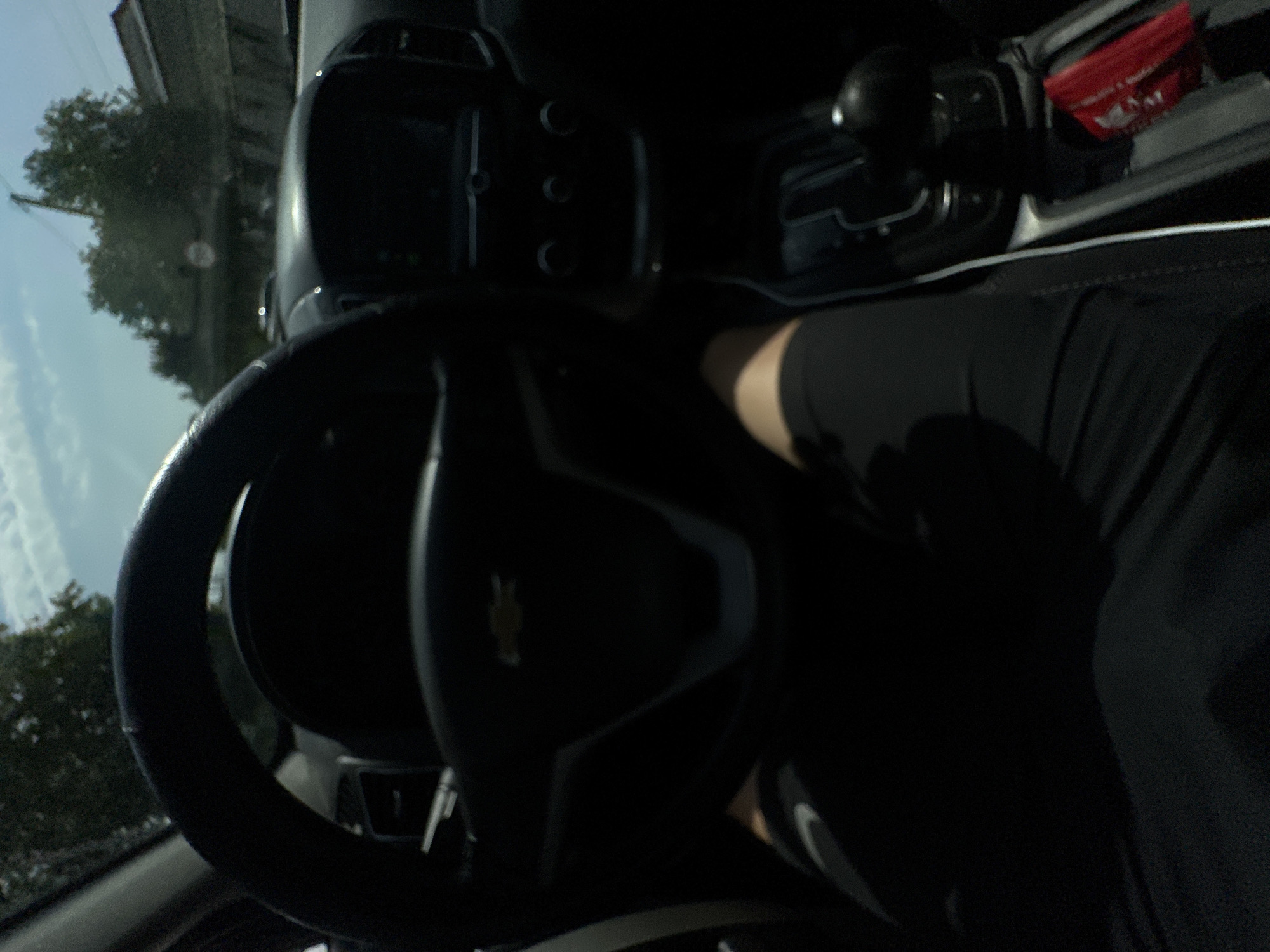 The image shows a first-person view from inside a car, looking at the steering wheel and dashboard. The car appears to be stationary, possibly parked or stopped at a traffic light, with the engine off. The exterior is visible through the windshield, showing trees, a building, and a clear sky, suggesting a daytime scene in a moderately urban or suburban environment.
The interior of the car is dark, with the steering wheel and central console being the most prominent features. A person's legs are visible in the lower part of the image, wearing black shorts. The steering wheel has a Chevrolet logo in the center. The gear shifter is visible, suggesting an automatic transmission. A red object, possibly a wallet or a packet of tissues, is on the center console.
Without specific visual cues in the image that are unique to Tashkent, Uzbekistan, it is impossible to confirm the location. However, the overall scene is consistent with a typical car interior in many parts of the world.