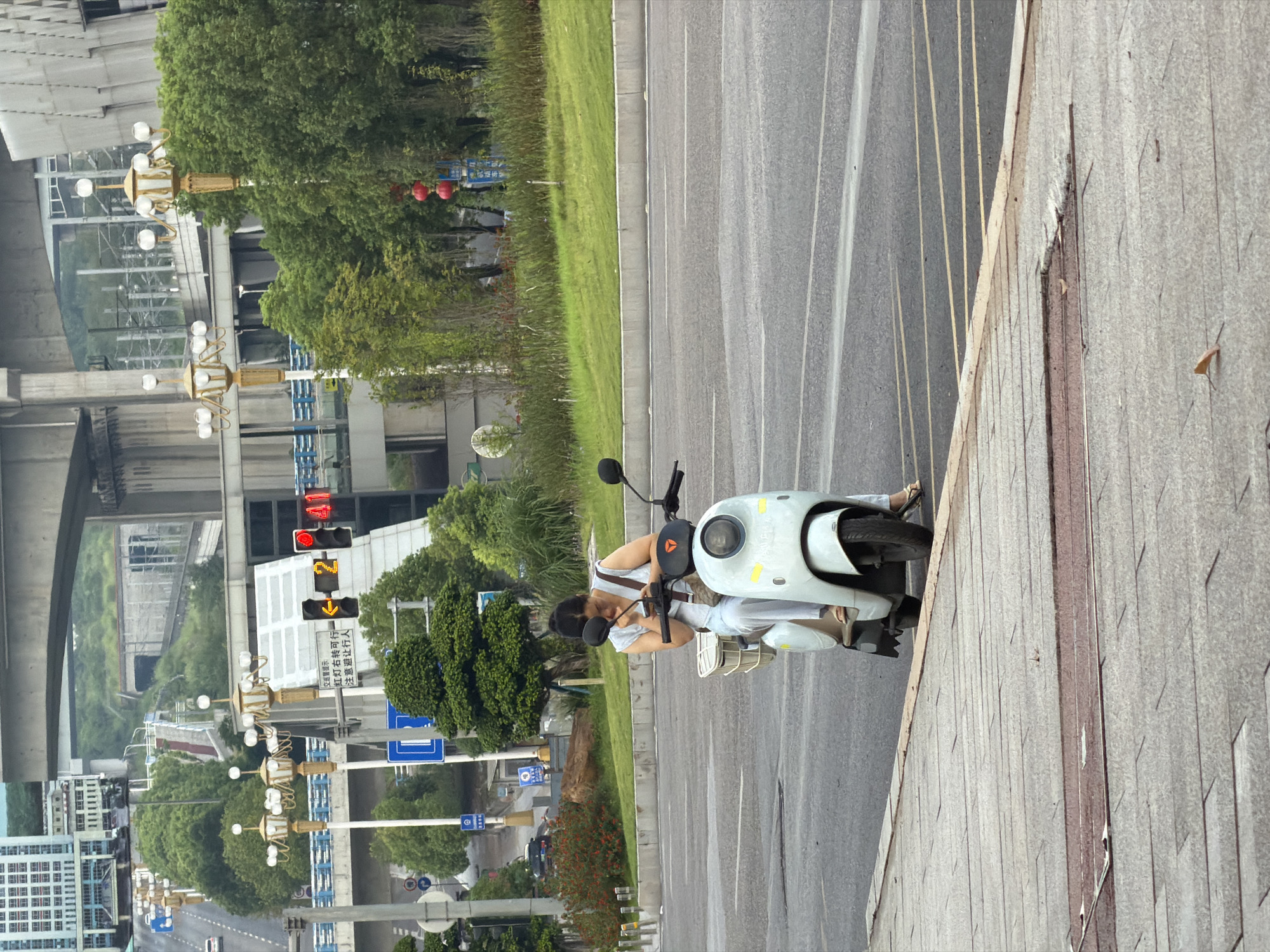 In Jiangjin, China, a woman sits on a white scooter at a traffic light, looking at her phone. She is wearing a light blue top and white pants. The scooter has a basket on the front. The background shows a multi-level highway overpass, lush green trees, and several ornate streetlights. Red lanterns are visible hanging from a pole on the right side of the image. The road is paved and marked with yellow lines. The sidewalk in the foreground is made of gray paving stones with some wear and tear.