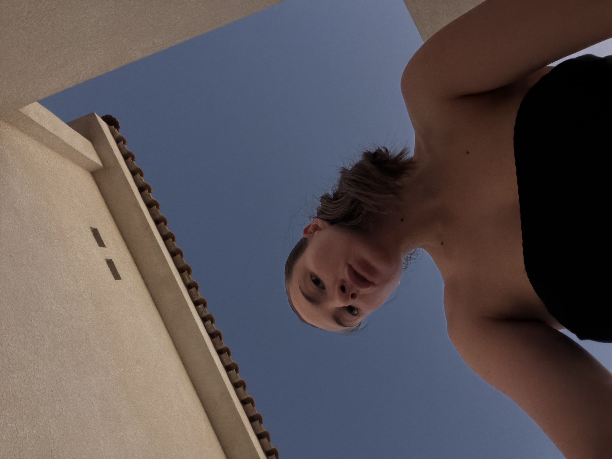 A low-angle medium shot captures a young woman from the shoulders up, looking directly at the camera with her head tilted slightly down. She is wearing a black strapless top. Her hair is pulled back into a loose bun. Behind her, the scene is dominated by a clear, deep blue sky. To the top left of the frame, a beige-colored awning or overhang with visible roof tiles is visible. To the right, a portion of a beige wall is present. The overall impression is of an outdoor setting, possibly a balcony or terrace, under a bright sky. There is no indication of Sharm El Sheikh, Egypt, in the image itself.