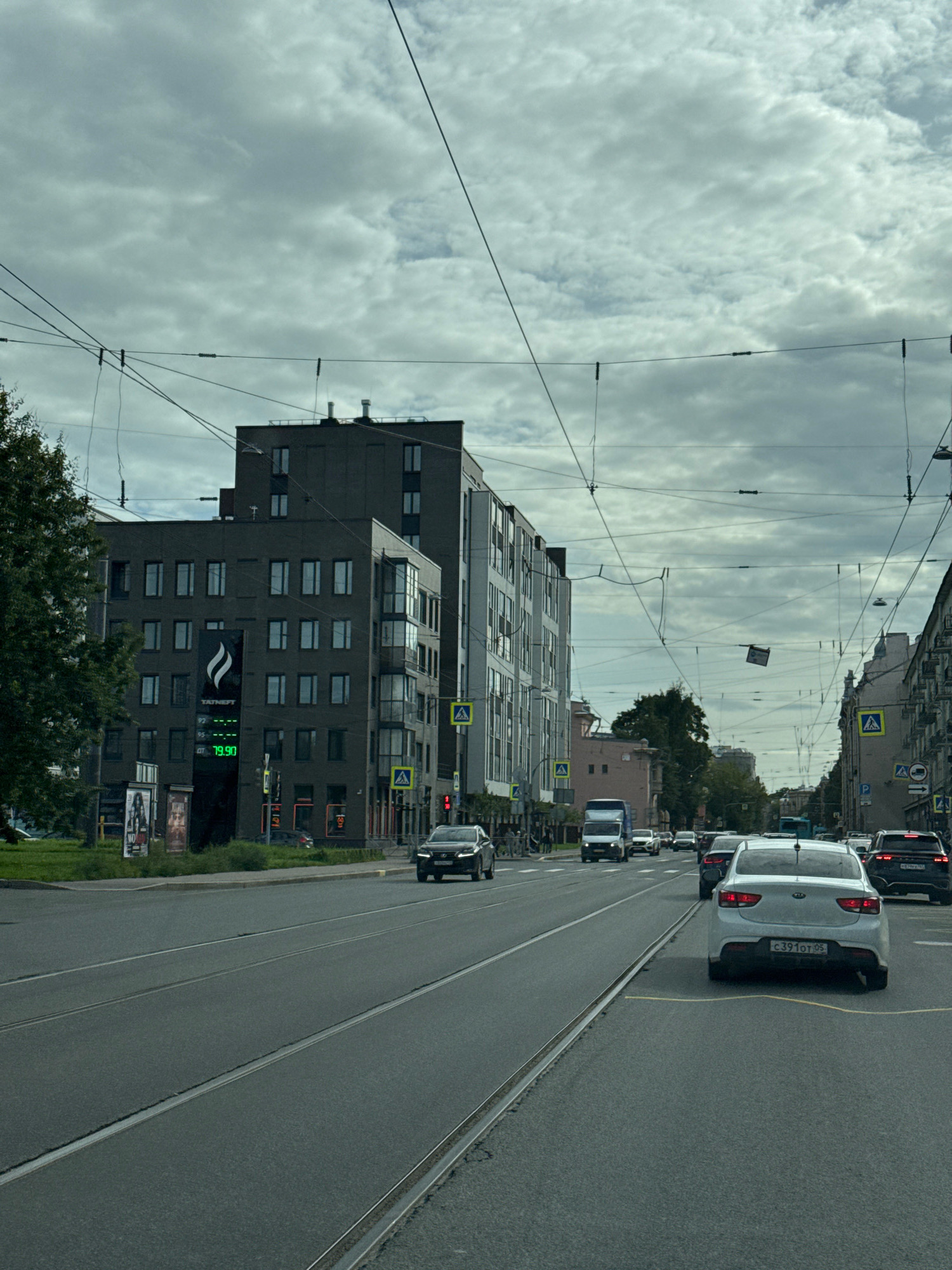 The image captures a street scene in Saint Petersburg, Russia, on a cloudy day. The perspective is from inside a vehicle moving along a wide, multi-lane road that features tram tracks. On the left side of the road, a modern, dark-colored building with numerous windows is prominent, displaying a large advertisement for Tatneft with a price of 79.90. Further down the street, more buildings, some with intricate architectural details, line both sides of the road. Several cars are visible, including a white Kia in the foreground with a license plate starting with C391OT. Traffic lights and pedestrian crossings are present. Overhead, a complex network of electrical wires for trams crisscrosses the sky, with a piece of paper or fabric hanging from one of them. Trees and greenery are visible on the median and sidewalks. The overall atmosphere is that of an urban environment with active traffic.