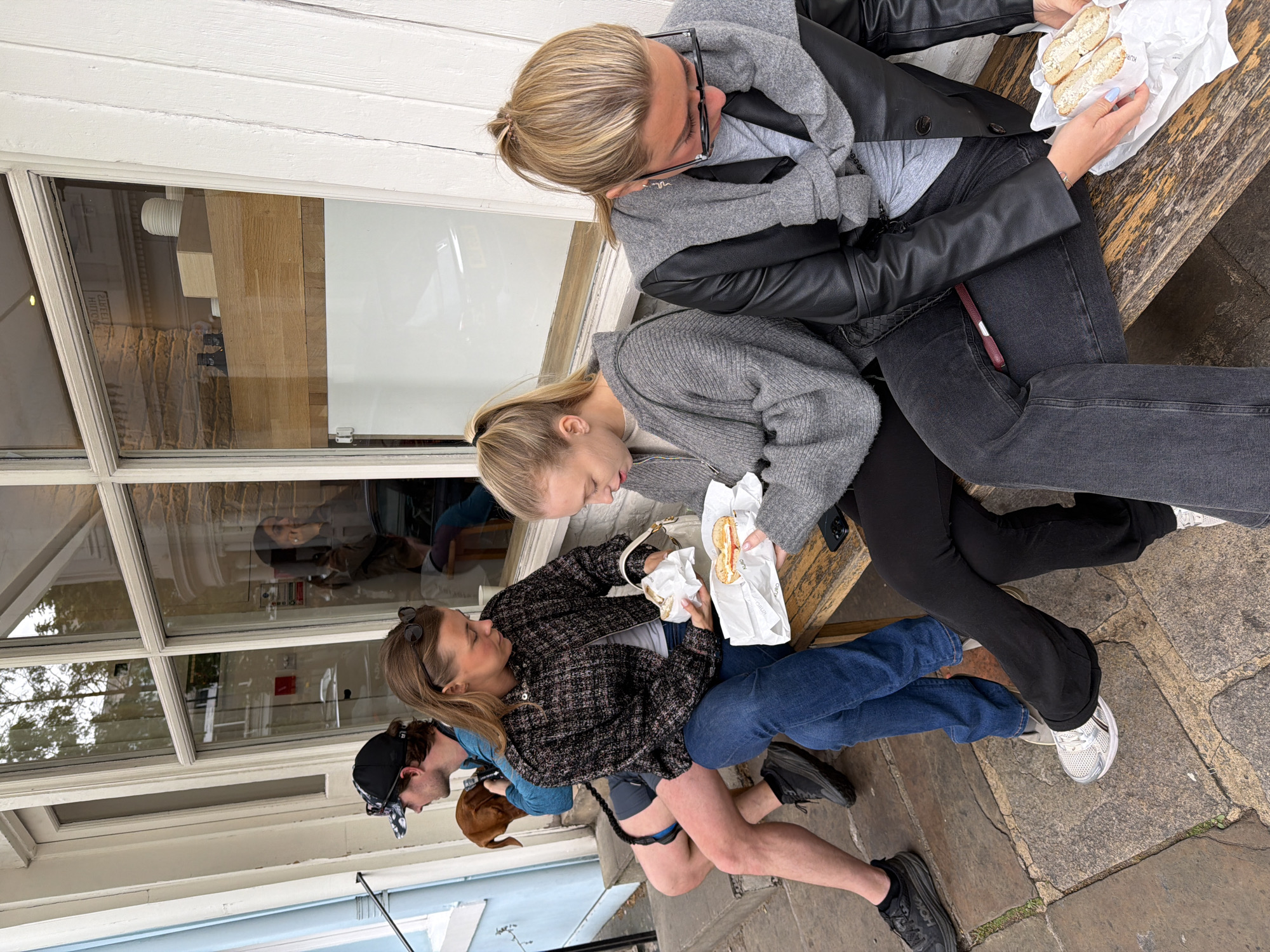 A horizontal, slightly angled eye-level shot shows five people sitting on a bench outside a building with large windows and white wooden frames, likely in London, United Kingdom. The upper two women are seated side-by-side. The woman on the left has blonde hair in a ponytail, wears glasses, a grey sweater, and a black leather jacket. She holds a white paper bag containing halves of a filled roll. The woman next to her has blonde hair in a ponytail and is wearing a grey cardigan. She also holds a white paper bag with a filled roll.
The next two people sit on the same bench, to the lower left. The woman is wearing a black-and-white jacket and blue jeans. The man beside her wears a backwards baseball cap, glasses, and a blue shirt. There is a small brown dog in his lap. A fifth person sits beside the man; they wear black shorts and a black T-shirt, with a harness around their thigh.
The building's windows reflect various objects and people inside. The ground in front of the bench appears to be paved with stone. The building's exterior is white with blue accents.