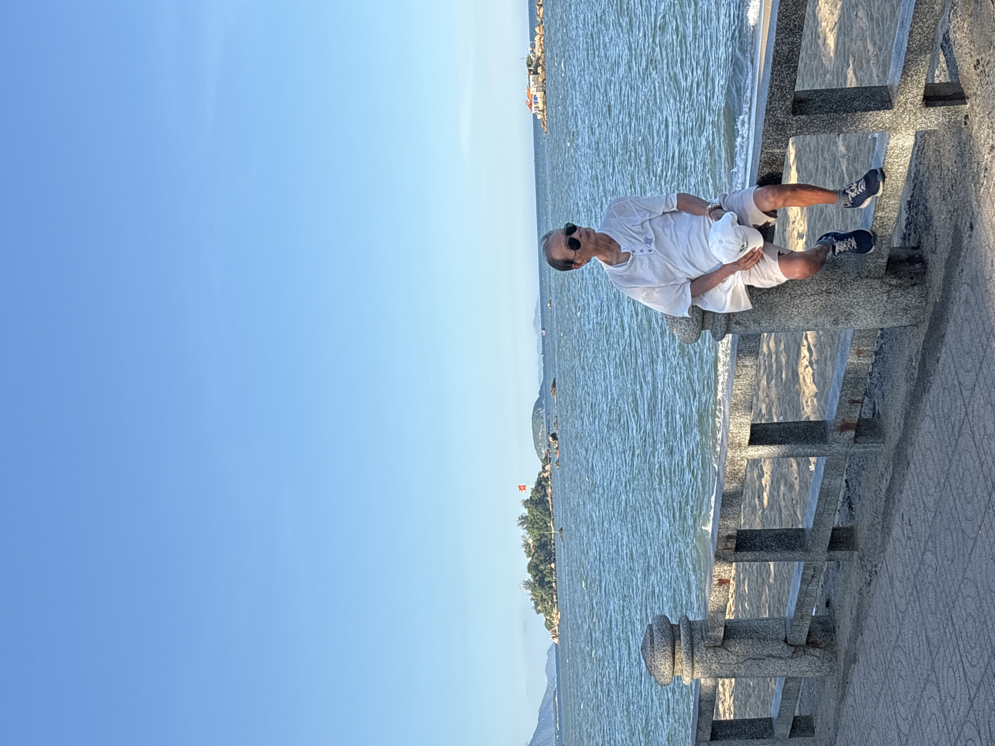 An elderly man wearing sunglasses, a white short-sleeved shirt, and light-colored shorts is sitting on a stone railing overlooking a beach and the ocean. He is holding a white cap in his lap. The railing is part of a promenade or walkway, made of stone with square openings. The beach behind the railing is sandy, and the ocean has small waves. In the distance, there are several small islands, one of which has a red flag flying. The sky is clear and blue. The location is likely a coastal area in Nha Trang Ward, Vietnam, given the presence of islands, the sea, and the tropical-looking vegetation on the islands.