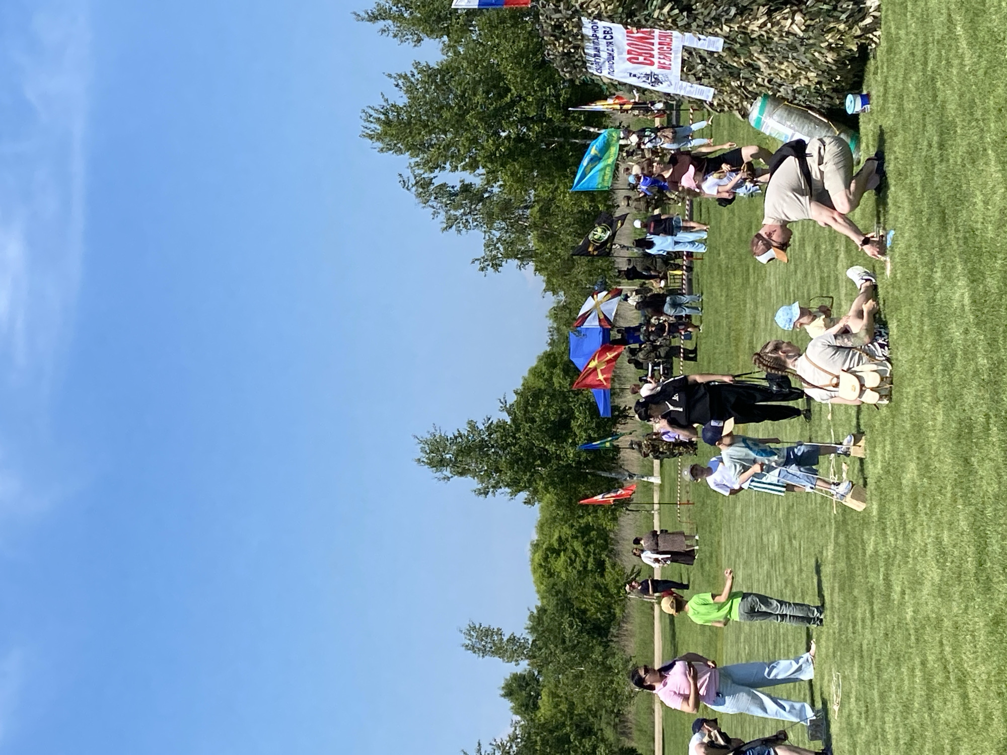 An outdoor event is taking place on a bright, sunny day on a grassy field, with a line of trees in the background under a clear blue sky. In the foreground, several families are gathered. On the left, a woman in a pink shirt and light blue pants stands and watches. To her right, a young boy in a green shirt and dark pants stands. Further right, two children are walking on stilts made of wood. In the middle ground, several people are standing and talking, some in military-style clothing. Flags of various countries are displayed, including Russia, Kazakhstan, and others. A camouflage-covered structure is visible on the right side of the image. In the lower right, a man in a tan shirt and shorts is crouching down, appearing to be showing something to two children sitting on the grass. One child is wearing a tan shirt and shorts, and the other is wearing a light blue hat. A plastic water bottle and a blue bucket are near the crouching man.
