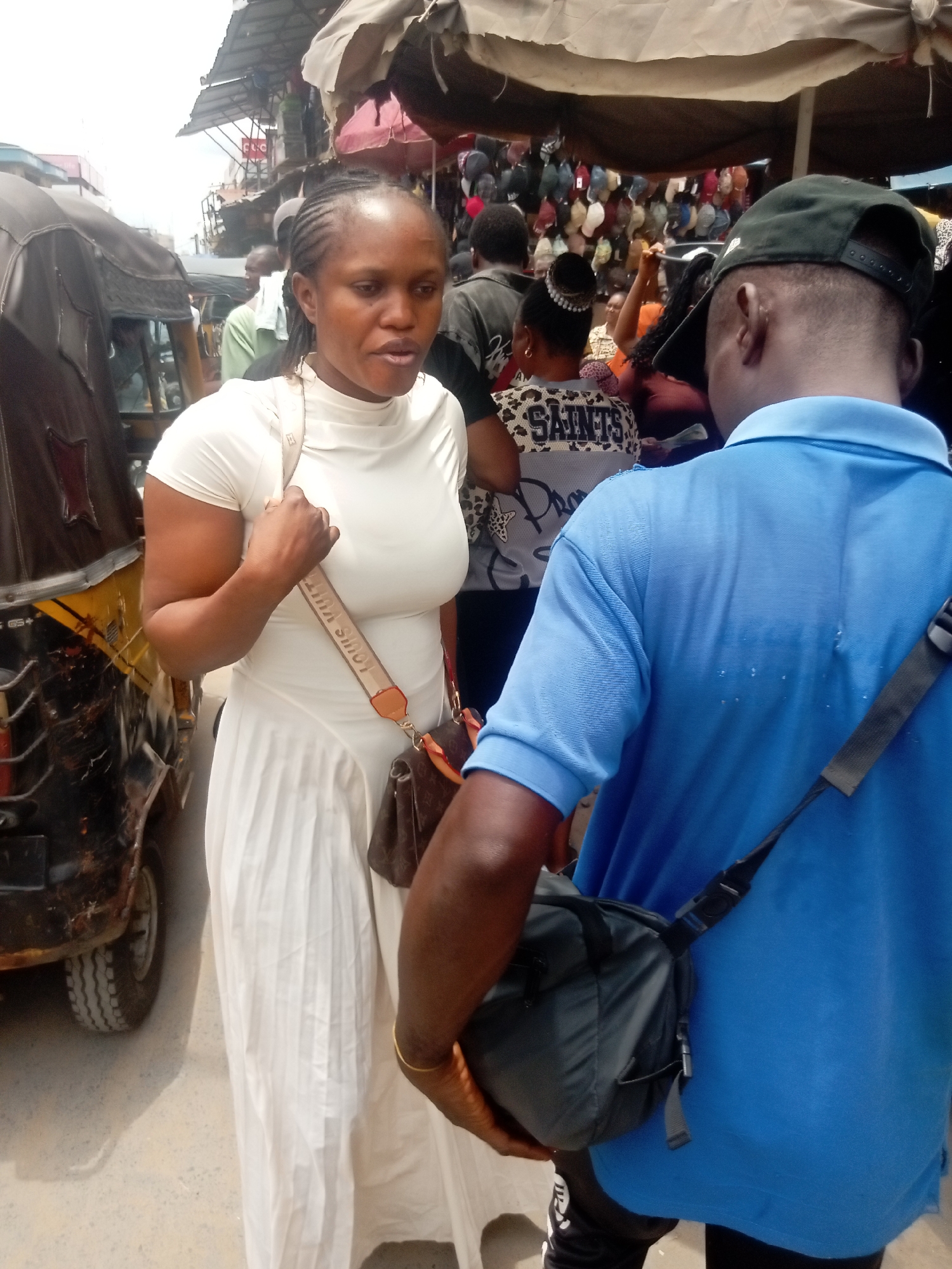 The image depicts a bustling street scene in Fegge, Nigeria. A woman in a white outfit with a Louis Vuitton bag is engaged in conversation with a man wearing a blue shirt and a black cap, who is holding a black bag. An auto rickshaw is parked nearby. In the background, a crowded market area is visible under a makeshift canopy. Vendors are selling hats. Other people walk through the street.