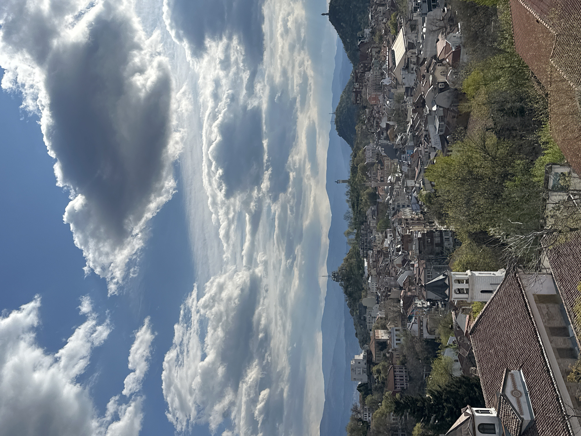 The image shows a cityscape under a partly cloudy sky. The cityscape consists of tightly packed buildings, mainly residential houses, with a variety of architectural styles and red tiled roofs. Green trees are interspersed among the buildings. In the background, there are hills and mountains. The sky is blue with many white cumulus clouds. The view appears to be taken from a high vantage point.
The location in the city of Plovdiv, Bulgaria, is suggested by the architecture and the hilly landscape. The presence of the historical buildings and the terrain are consistent with the city's geography.
