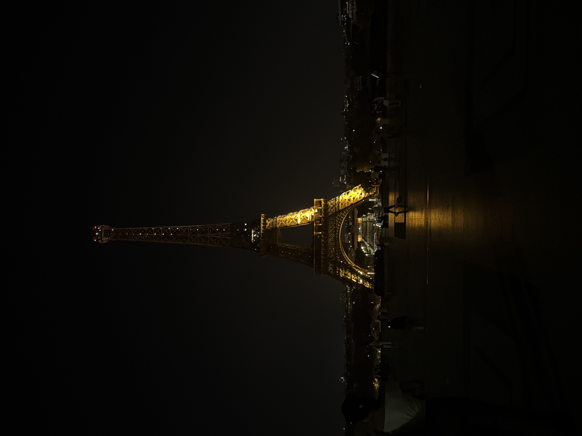 The image depicts the Eiffel Tower at night in Paris, France. The tower is brightly lit, contrasting with the dark sky. A road and river (Seine) run alongside the tower, partially illuminated by streetlights and reflections. People are present, but their features are not clear. The image is oriented sideways.