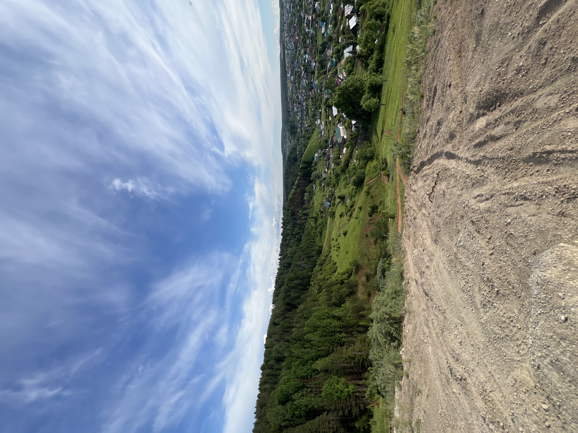 The image, taken from a high vantage point, presents a sweeping landscape view of a rural area, likely Ольховка, Russia. The upper half of the frame is dominated by a clear blue sky, adorned with scattered, wispy white clouds that stretch across the expanse.
In the middle ground, a lush, dense forest of green trees covers a significant portion of the scene, suggesting a hilly or sloped terrain. Beyond the forest, on a higher elevation, a sprawling village or small town is visible, with numerous houses featuring diverse colored roofs (such as green, red, and gray), indicating human settlement nestled within the natural environment.
To the right in the foreground, a large, barren dirt and gravel slope descends sharply. This area shows signs of erosion or disturbance, with visible tracks or patterns etched into the light brown earth. A narrow, unpaved path can be seen winding through the grassy sections between the dirt slope and the forest, leading towards the distant village.
The photograph captures a bright, sunny daytime scene, with clear weather and no specific activities or people immediately apparent. The overall impression is one of serene, expansive natural beauty alongside rural development. There is no visible text in the image.