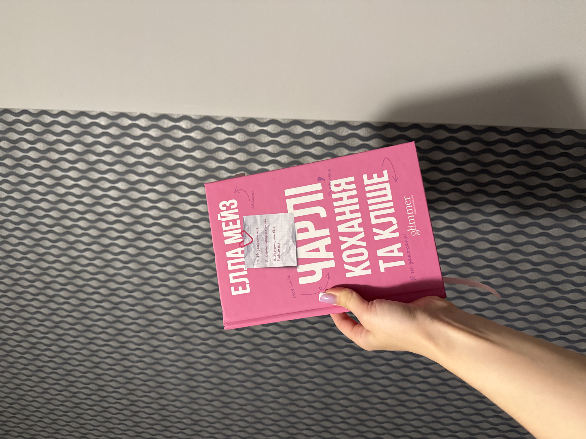 A person's hand with a pink-purple fingernail polish is holding a bright pink book titled "ЧАРЛІ КОХАННЯ ТА КЛІЩЕ" (Charlie Love and the Tick). The book has a handwritten note attached to the cover with a small heart drawing. The note reads: "1. Не закохатися. 2. Бути професійкою. 3. Забути, як він дивиться..." (1. Don't fall in love. 2. Be professional. 3. Forget how he looks...). Another note on the book says "Моє ім'я" (My name), and a smaller note below the title says "Не про мене" (Not about me). The book also has a logo for "glimmer PUBLISHING HOUSE". The background of the image consists of a textured gray wall with a repeating wavy pattern. There is no information in the image to suggest a specific location in Łódź, Poland.