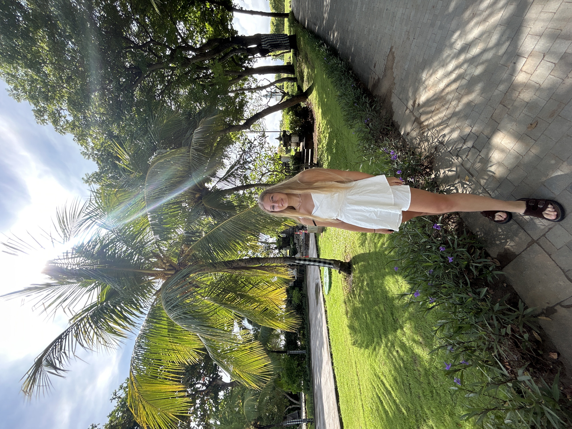 The image displays a full-length shot of a young woman standing outdoors in a lush, tropical environment, consistent with the provided location context of Kuta Selatan, Indonesia. The woman has long blonde hair and is wearing a white short dress and dark sandals, smiling faintly towards the viewer.
She is positioned on a vibrant green lawn next to a paved pathway that recedes into the background. A large palm tree with visible coconuts is prominent in the foreground to her left, its fronds illuminated by bright sunlight which also creates a noticeable lens flare. Beyond her and the palm tree, numerous other large trees with dense foliage cast dappled shadows. A traditional Balinese-style stone structure or shrine is visible amongst the trees in the mid-ground. One of the tree trunks in the background is adorned with a black and white checkered cloth, a common cultural signifier in Bali.
The scene is brightly lit by the sun, suggesting daytime, either morning or late afternoon, with clear skies and some scattered clouds. Strong shadows of leaves and trees are cast across the paved pathway and the grassy area. A small border of green plants with scattered purple flowers lines the edge of the pathway and lawn. The overall atmosphere conveys a sense of peace and a relaxed, sunny vacation setting. No text is visible in the image.