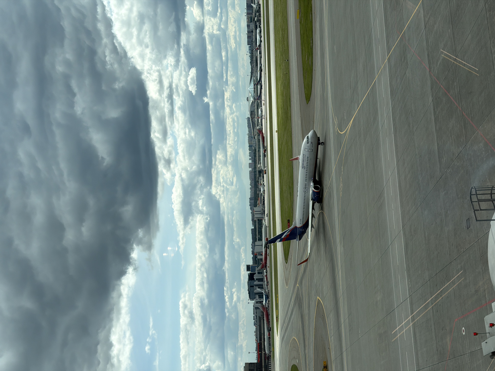 The image depicts an Aeroflot airplane on a runway at Sheremetyevo International Airport in Khimki, Russia. The view is from an elevated perspective, possibly from an airport terminal window. The sky is partially overcast with dark, heavy clouds contrasting with patches of blue sky and white clouds. Other aircraft are visible parked along the runway, and there are some buildings in the background. The runway is marked with yellow and white lines.