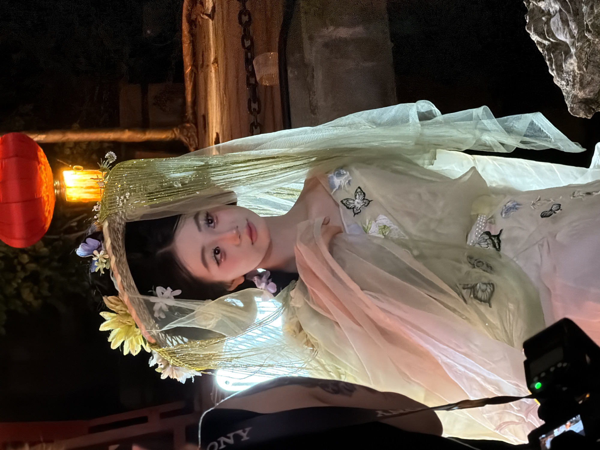 A young woman wearing traditional Chinese attire stands outdoors at night. She is adorned with a headpiece featuring flowers and a veil, and her dress is light green with floral and butterfly embroidery. A red lantern hangs in the background, illuminating the scene. The woman is looking to the right, with her makeup emphasizing her eyes and cheeks. In the foreground, a camera is visible. The setting appears to be a street or park in Yuzhong District, China, indicated by the presence of a chained fence and stone elements.