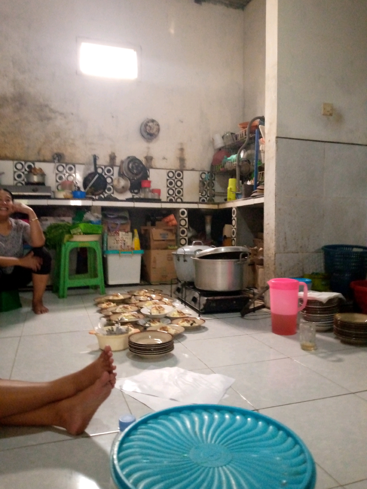 The image depicts the interior of a kitchen or dining area in Sedati, Indonesia, characterized by tiled flooring and partially finished or rustic wall plaster. A woman is seated on a green stool in the left foreground, facing left and smiling. Her left arm is resting on her head. In front of her, a pile of plates with food is laid out on the floor, suggesting a communal meal preparation or a recent gathering. Various kitchen appliances and utensils are visible, including pots, pans, a stove, and shelves stocked with containers and dishes. A large, blue plastic tray is in the bottom right of the frame, and a pink pitcher is on a counter in the middle ground. The overall ambiance suggests a busy, domestic setting.