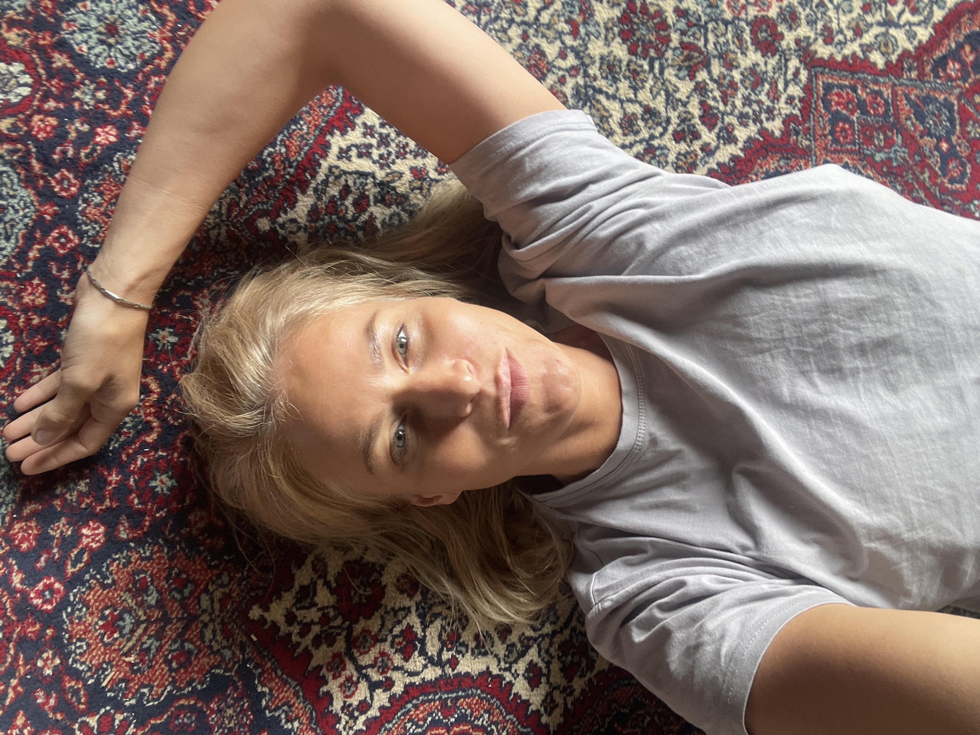 A blonde woman wearing a gray t-shirt is lying on a patterned rug. She is facing the camera with her head resting on the rug. Her left arm is extended and slightly bent at the elbow. Her right arm is bent at the elbow and extends towards the lower right of the frame. The rug has a complex pattern in red, blue, and white. The location in the city of Baranovka, Russia is not discernible from the visual information in this image.