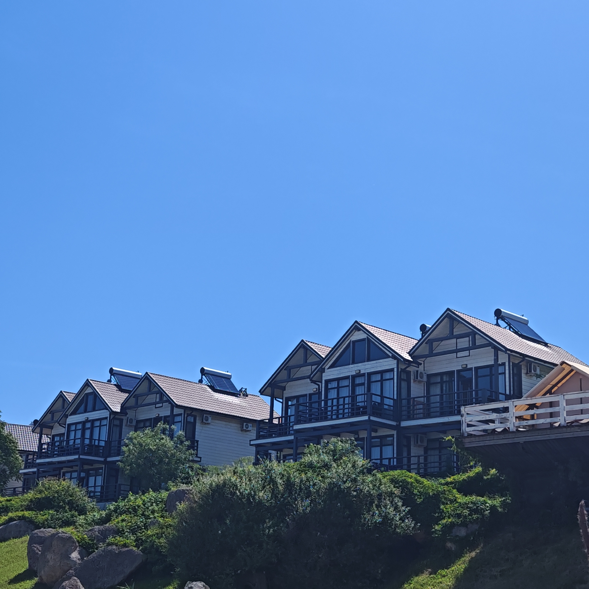 A daytime outdoor photograph depicts a row of at least five multi-story residential structures built into a green, rocky hillside under a clear, cloudless, cerulean blue sky. The scene is devoid of human presence.
The houses, consistent in architectural style, exhibit light-colored (appears off-white or cream) exterior walls, contrasted with dark brown or black gabled roofs topped with terracotta-colored tiles. Each visible residence features multiple balconies with dark railings on at least two levels, equipped with large, dark-tinted windows and glass doors. Solar water heaters, identifiable as cylindrical tanks resting on tilted dark panels, are installed on the roofs of at least three of the houses. White rectangular air conditioning units are visible mounted on the exterior walls beneath some balconies. On the far right, a segment of a white wooden railing of an elevated structure, possibly a deck or connecting walkway, is partially visible.
The terrain below the structures is characterized by numerous large, dark grey boulders or rocks interspersed with lush, dense green shrubbery, bushes, and patches of grass. The sunlight is bright and direct, illuminating the scene evenly. The image depicts a location within Мысовское сельское поселение, Ukraine.