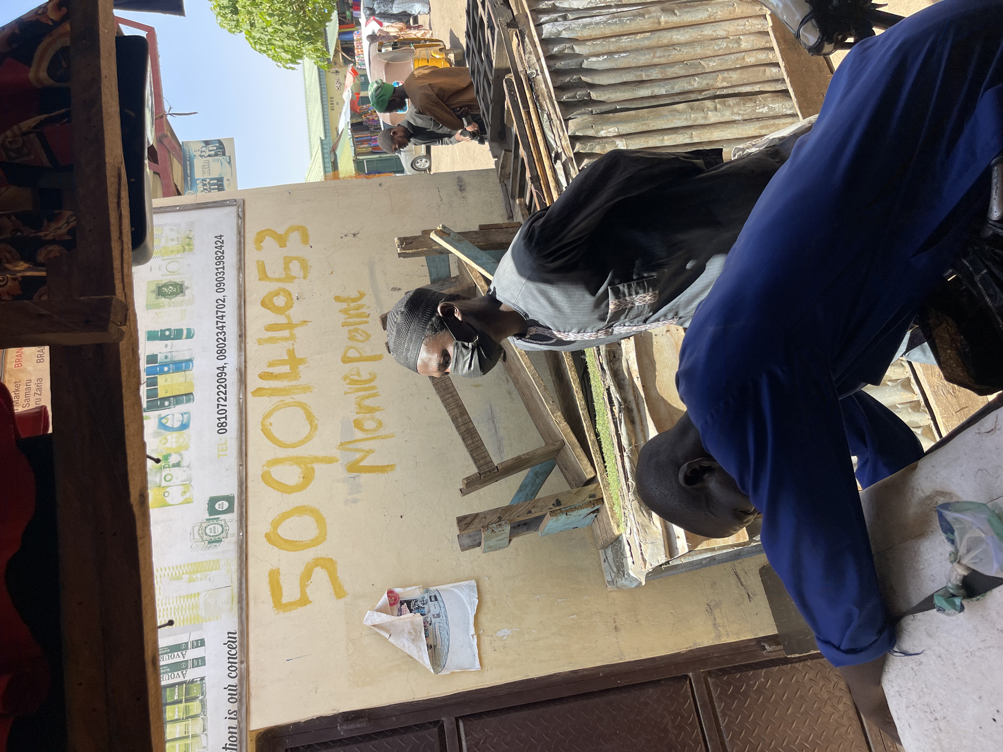 The image shows a street scene in Samaru, Nigeria. Two men are visible; one wears a black shirt and cap with a face mask, and another wears a blue shirt. The first man is facing the camera. In the background, another man wearing a brown shirt and green cap is visible. The scene appears to be a commercial area, with a sign displaying the phone number "5090144053" and the words "Monie Point" painted on a wall. The sign also contains phone numbers "08107222094, 08023474702, 09031982424." A sign reading "Market Samaru Zaria" is visible on the side of a building. A discarded piece of paper is affixed to the wall near a wooden structure and a corrugated metal awning.