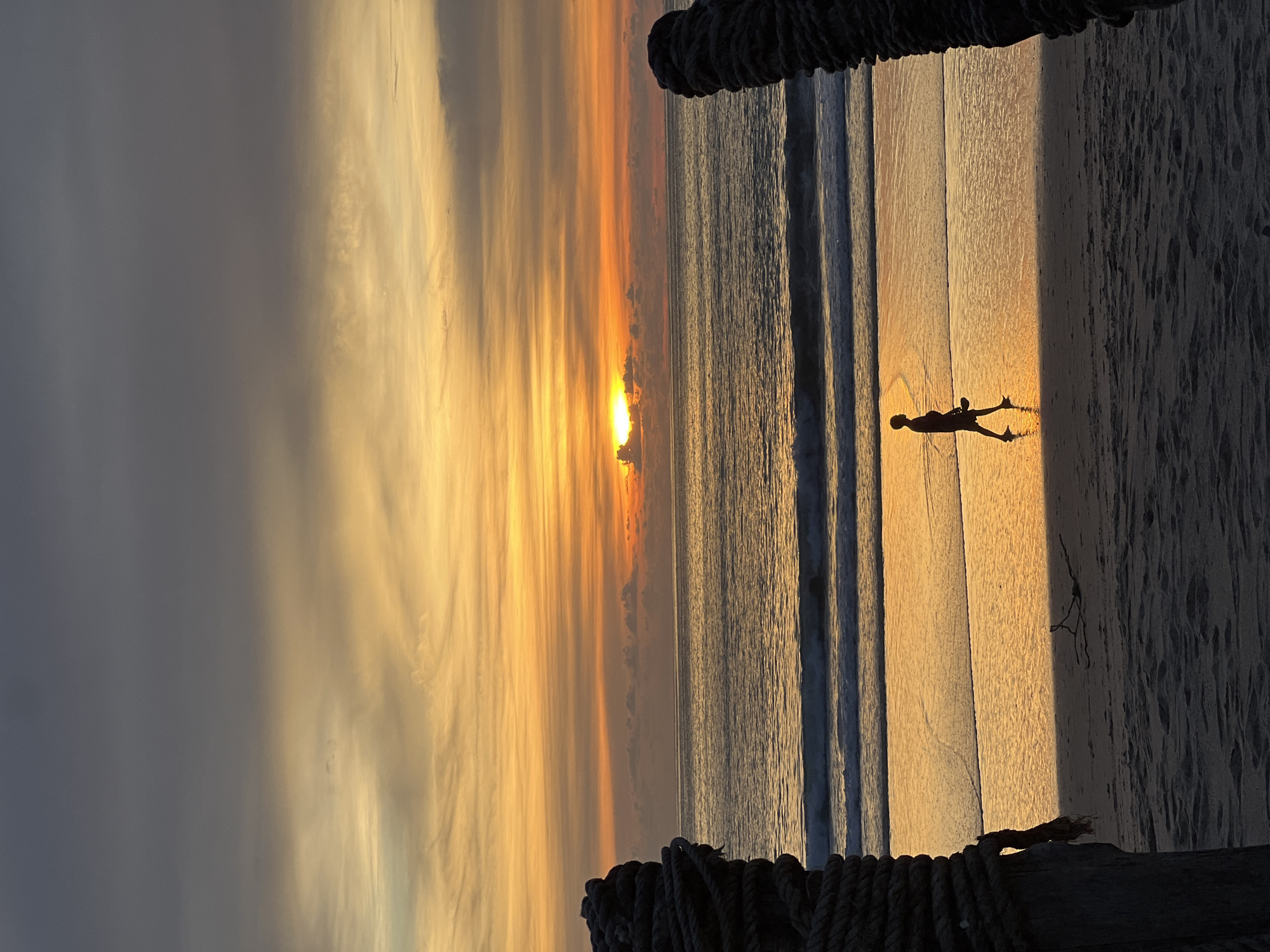 A lone figure walks along a wet beach as the sun sets over the ocean. The sky is a gradient of colors, with dark gray clouds at the top transitioning to bright orange and yellow hues near the horizon. The sun, partially obscured by clouds, casts a warm glow on the water and sand. Two large, rope-wrapped posts frame the scene on either side, suggesting a pier or some form of beachside structure. The figure, silhouetted against the sunset, appears to be holding something in their hand and is walking towards the right of the frame. Waves gently lap at the shore, and the wet sand reflects the colors of the sky. The overall atmosphere is serene and peaceful.
