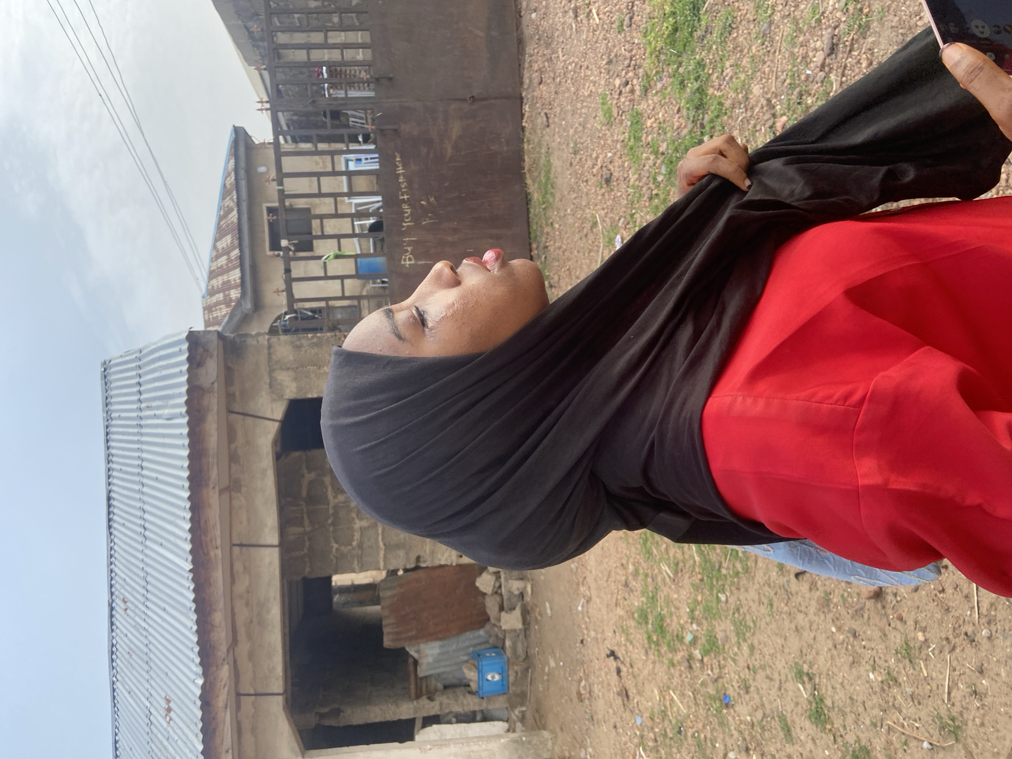 A medium shot captures a person, likely a woman, from the chest up, looking upwards and slightly to the right of the frame. She has medium to dark skin, red lipstick, and is wearing a red garment under a black head covering or scarf, which she holds up with one hand. Her left hand is visible on the right side of the image, holding a smartphone with emojis visible on its screen.
The setting is outdoors, possibly a semi-urban or rural area, under a bright, clear daytime sky. In the background, there are several simple buildings with unpainted concrete block walls and corrugated metal roofs. One building features a tall, dark metal gate, on which faint white text is visible, reading "BUY YOUR FISH HERE". The ground is unpaved, consisting of dirt and sparse patches of grass, with some debris like small rocks and sticks. A blue plastic crate is partially visible near the base of one building.
