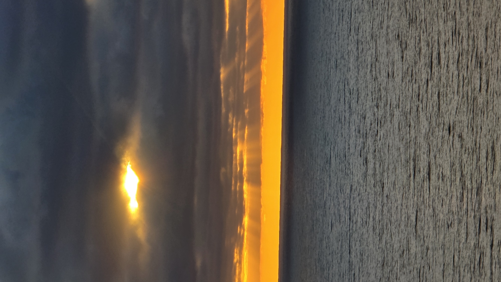 The image shows a dramatic sunset over the Pacific Ocean. Dark, heavy clouds dominate the upper portion of the sky, with a bright, golden light breaking through a gap in the clouds, casting rays downwards. Below the clouds, a vibrant band of orange and yellow stretches across the horizon, illuminated by the setting sun. The ocean is a deep blue-grey, with a rippled surface reflecting the colors of the sky. The scene appears to be captured from a high vantage point, possibly a cliff or bluff overlooking the coastline in Encinitas, California. There are no people, subjects, or objects visible in the frame, only the natural elements of sky and sea.