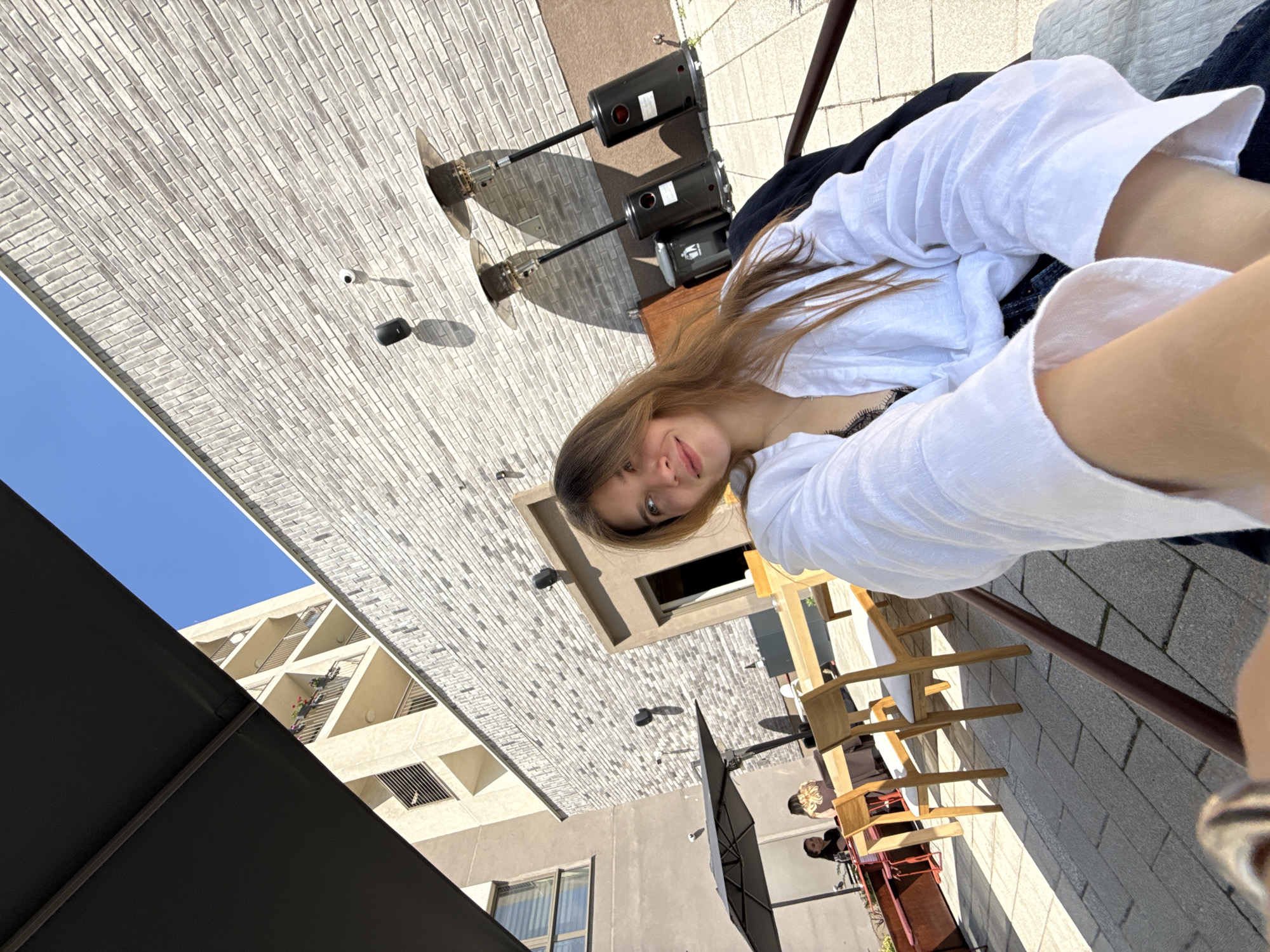 A young woman with light brown hair wearing a white shirt is taking a selfie while seemingly sitting outdoors. In the background, there is a clear blue sky visible next to a light-colored brick building. To the right of the woman, outdoor heaters, tables, chairs, and people are visible in an outdoor dining area. Further behind the woman and dining area is an outdoor sun umbrella. Based on the architectural details and street furniture, this image appears to be taken in the city of Belgrade, Serbia.