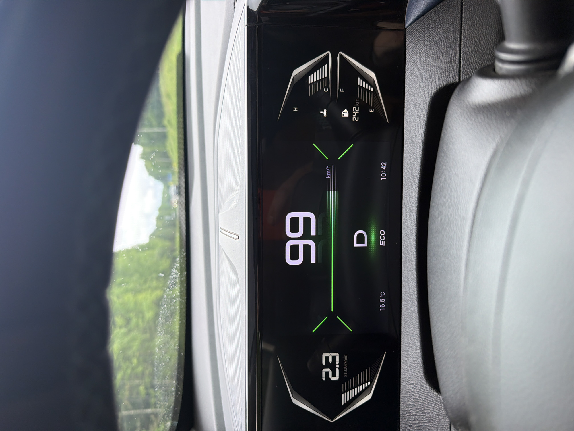 The image displays the interior of a vehicle, specifically a digital instrument cluster and a view through the windshield, depicting a scene in the city of Garevaya, Russia. The car is in motion, evidenced by the speedometer showing "99 km/h" and the tachometer indicating "2.3 x1000 r/min." The transmission is in "D" (Drive) mode, with "ECO" efficiency mode activated. The ambient temperature is displayed as "16.5 °C," and the time is "10:42."
The instrument cluster features a modern design with prominent white numerical readouts against a dark background, accented by green lines and angular metallic-look graphics. On the right side, a coolant temperature gauge shows the level near "C" (cold), and a fuel gauge indicates approximately three-quarters full, with a remaining range of "242 km." The upper portion of a dark-colored steering wheel is visible at the bottom of the frame. The dashboard features textured surfaces, including a ridged silver-grey panel above the display and a pebbled dark-grey surface below.
Through the windshield, a multi-lane road or highway is visible. The external environment is characterized by dense green foliage, primarily trees and bushes, lining both sides of the road. The windshield exhibits numerous small water droplets, indicating rain. In the distance, other vehicles can be faintly seen on the road, along with indistinct road signs. The sky appears overcast or partly cloudy, suggesting daylight conditions.