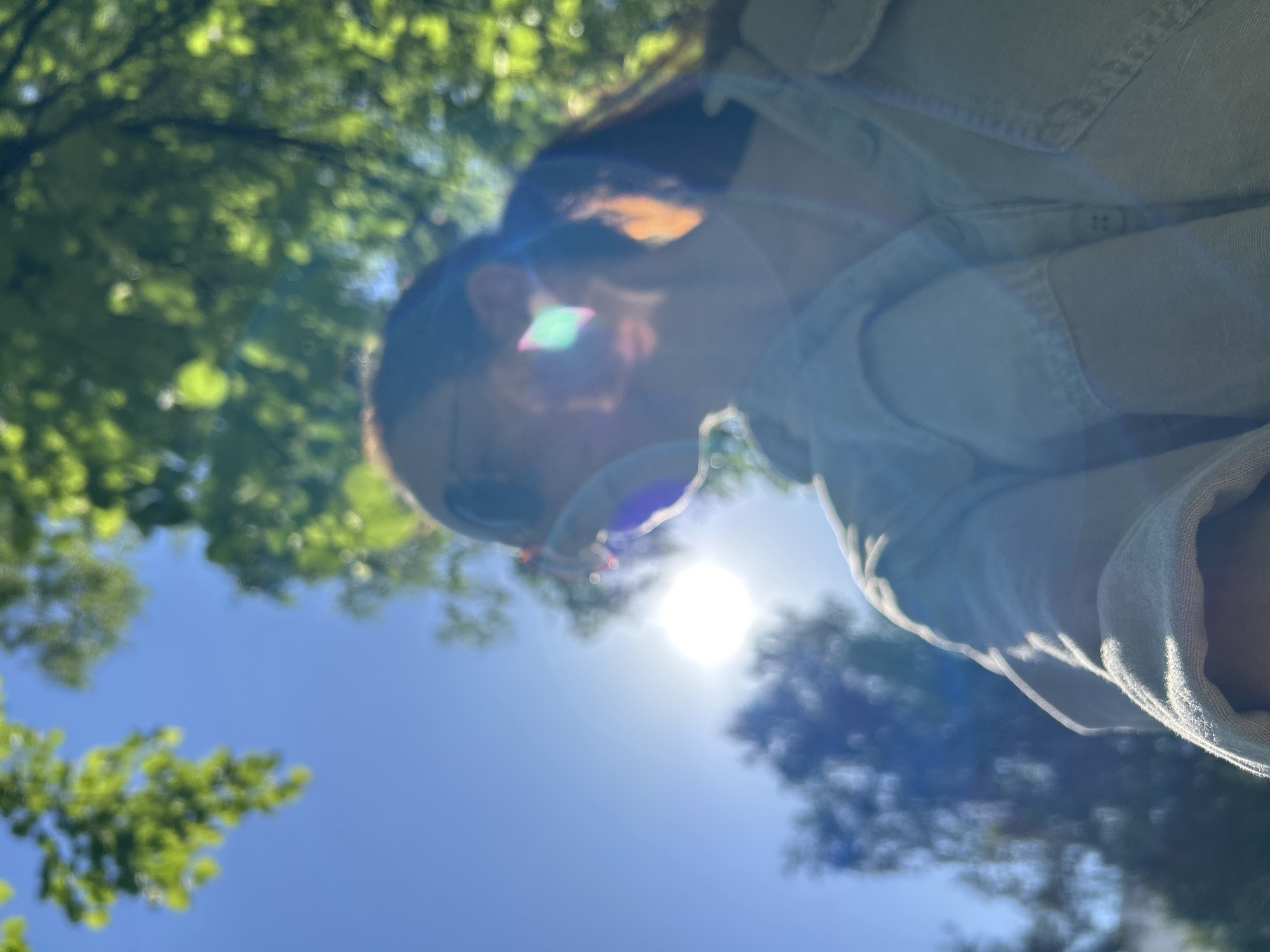 The image shows a low-angle view of a person in an outdoor setting, likely a park or wooded area. The person appears to be wearing a light-colored shirt or jacket and is facing downwards. The sun is directly overhead, creating a strong glare and lens flare. Trees and blue sky are visible in the background. The location, Chaikovsky, Russia, cannot be confirmed from the image.