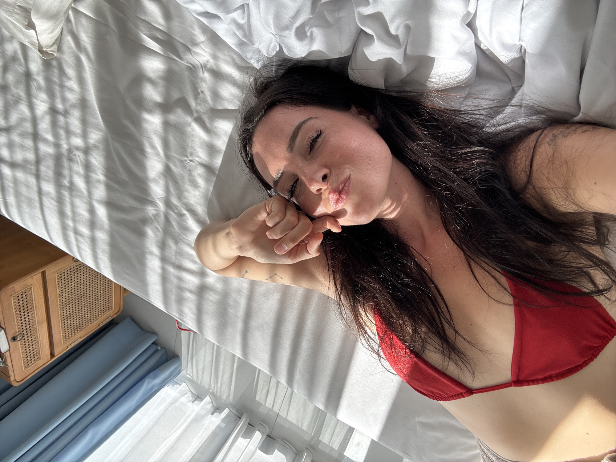 A high-angle, medium shot shows a woman lying on a bed, with sunlight streaming through blinds creating striped shadows on the white sheets. She is looking up at the camera, her head tilted to the side, with her right arm bent and her hand resting on her cheek. She is wearing a red bikini top. Her dark hair is spread across the pillow and her shoulder. To the left of the bed, blue curtains are visible, and beyond them, a light-colored wall. To the right of the bed is a bedside table with a woven drawer front and a small object on top. The location is identified as Mengwi, Indonesia.