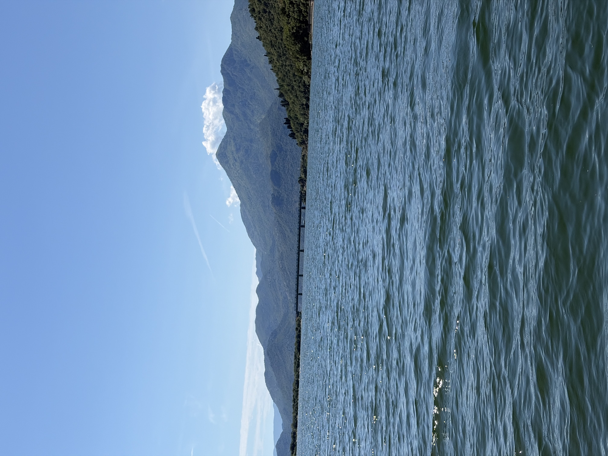 The image displays a wide-angle landscape featuring a large body of water, presumed to be a lake, situated in the area of Kruševica, Montenegro. The foreground and mid-ground are occupied by the water, which exhibits a dark blue-green hue with discernible ripples and small waves. Numerous small, bright reflections of sunlight are visible across the water's surface, indicating clear, sunny conditions. In the mid-ground, a long, dark-colored bridge, characterized by multiple spans, crosses the water, connecting forested landmasses. The background is dominated by a range of mountains, which are densely covered in green vegetation. Several mountain peaks are visible, with a notable white cumulus cloud positioned directly above one of the central peaks. The sky overhead is a uniform light blue, featuring a few faint, white contrails. No individuals, animals, or specific human activities are observable within the frame.