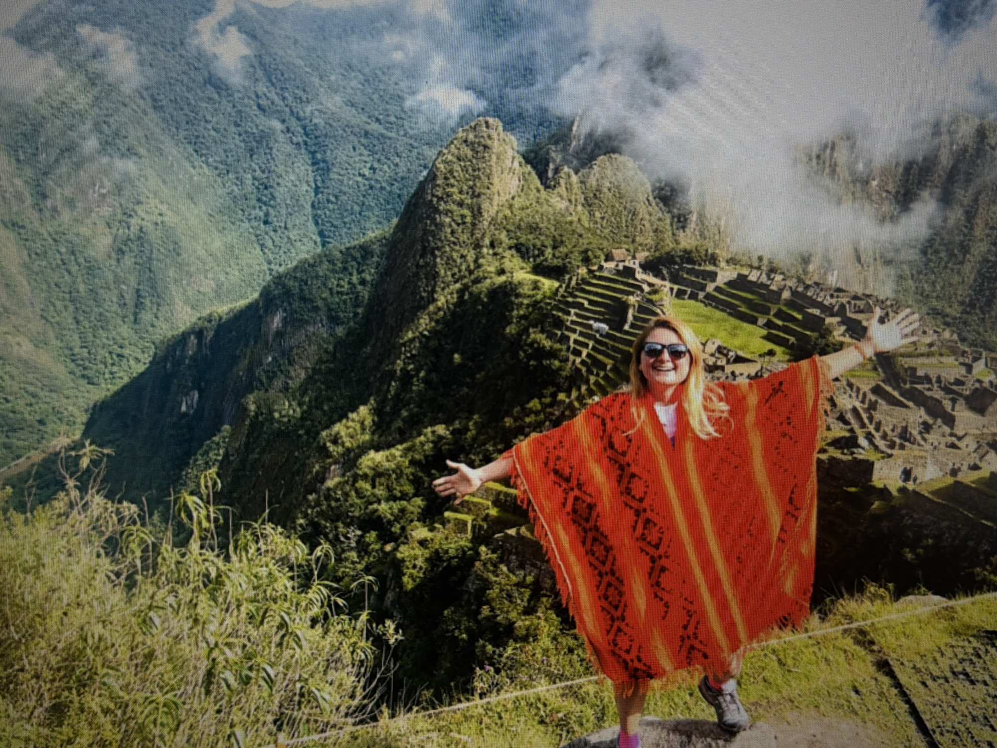 The image shows a woman standing with her arms outstretched in front of the ancient Inca site of Machu Picchu in Peru. She is wearing sunglasses and an orange poncho with a black patterned design. Behind her, the landscape includes mountains, ruins, and terraced fields. The weather is cloudy with some fog obscuring the mountain peaks. The foreground includes greenery and vegetation. The location is NOT in Krasnoyarsk, Russia, but at Machu Picchu in Peru.