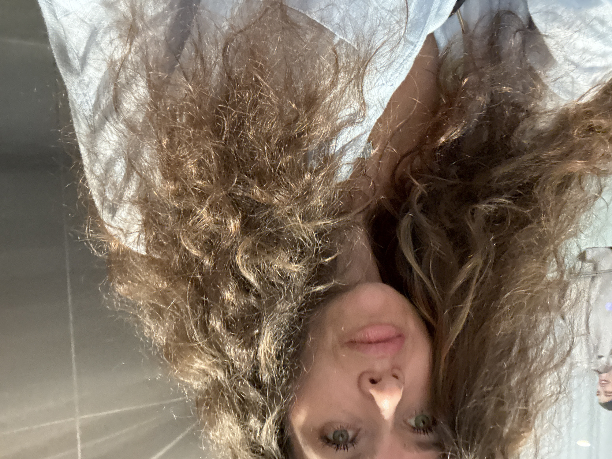A woman with long, wavy brown hair and green eyes takes a selfie in what appears to be an elevator in Dubai, United Arab Emirates. She is wearing a light blue shirt. In the background, a blurred image of another person is visible through a glass partition, suggesting the selfie is being taken in a public space. The elevator walls are metallic and have a grid-like pattern, consistent with elevator interiors found in modern buildings in Dubai.