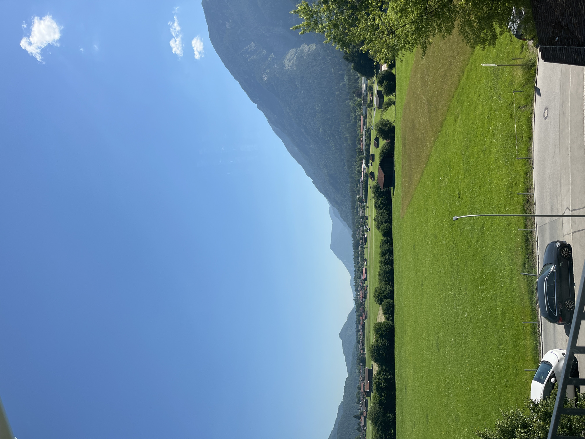 The image shows a panoramic view of a rural landscape with a village nestled in a valley between rolling mountains. The sky is a clear, bright blue with a few wispy white clouds. In the foreground, a lush green meadow stretches out, bordered by a line of trees. Several small, rustic buildings with red roofs are scattered across the meadow and in the village beyond. The mountains in the background are covered in dense green forests, their slopes rising majestically towards the sky. The scene appears to be taken from a slightly elevated vantage point, possibly a balcony or window, looking out over the picturesque countryside. The presence of cars on a paved road in the lower part of the image suggests it's a populated area, despite the rural feel. The overall impression is one of tranquility and natural beauty.