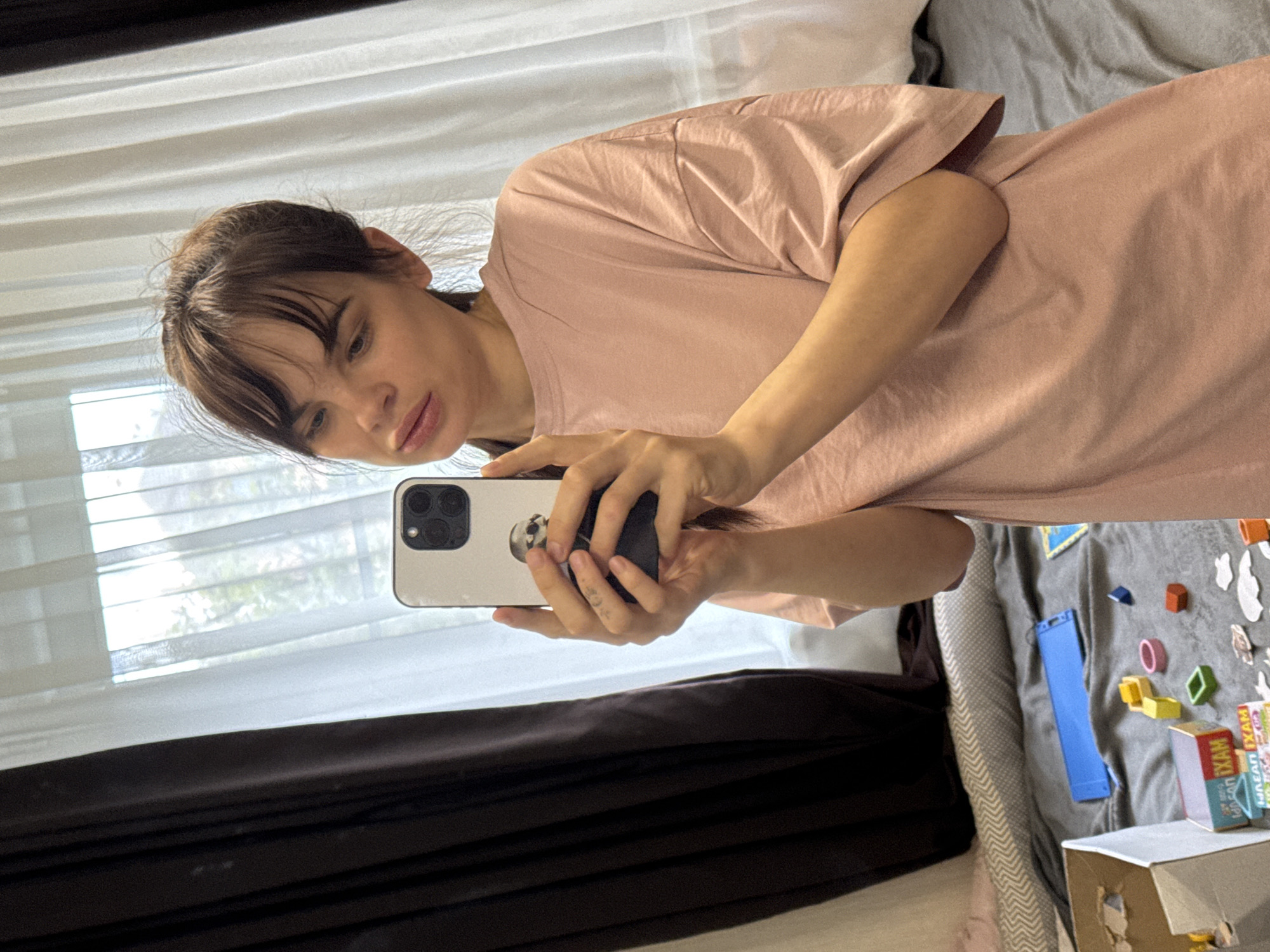 The image captures a young adult woman taking a mirror selfie indoors, rotated approximately 90 degrees counter-clockwise. She holds a light-colored smartphone, likely an iPhone due to its triple-lens camera system, in both hands. The phone case is light-colored and has a dark circular PopSocket featuring a skull design. The woman has brown hair with bangs, pulled back, and wears a loose-fitting, light pink (or peach) short-sleeved t-shirt. Her expression is neutral. Small dark markings, possibly a tattoo, are visible on one of her fingers.
The background suggests a domestic setting, possibly a bedroom or living area. To the left, a window is visible, covered by horizontal white sheer blinds or curtains, allowing natural daylight to illuminate the scene. Below the window are darker, solid curtains, appearing brown or black. To the right, a gray patterned bedspread or play mat is spread out, covered with various small, colorful children's toys and building blocks. These include yellow squares, orange and pink rings, green diamond shapes, and a long blue rectangular piece. Several small white cut-outs are also scattered on the mat.
At the bottom right, two cardboard boxes are partially visible. One small package prominently displays the word "EXAM" in black letters on a vibrant red, yellow, and blue background. Above it, part of the Cyrillic word "УМНАЯ" (UMNAYA, meaning "smart") is visible. The overall scene conveys a casual, everyday moment in Rostov-on-Don, Russia.