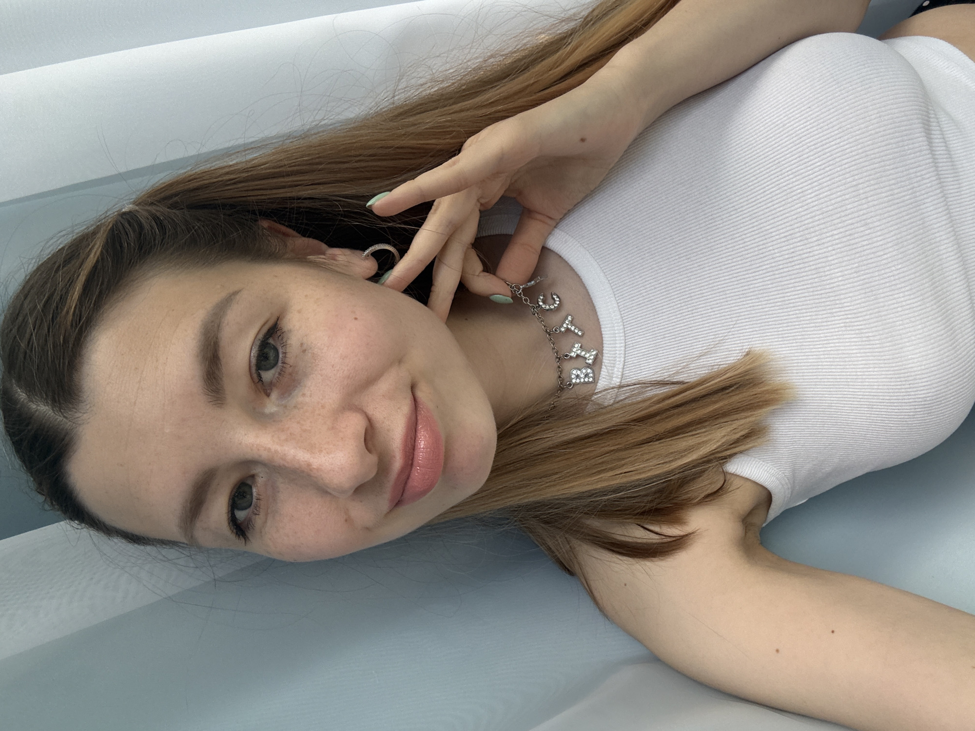 A young woman is lying down, likely on a light blue surface such as a bed or a similar structure. She is wearing a white tank top. Her hair is light brown, and she is wearing a necklace with the letters "CUTIE" in rhinestones. The woman has light skin, freckles on her face, and is wearing light pink lipstick. Her fingers are touching the necklace and a small hoop earring is visible in her ear. The background is a light, neutral color. The image appears to have been taken indoors with soft lighting. There is no information to suggest the location is in Moscow, Russia.