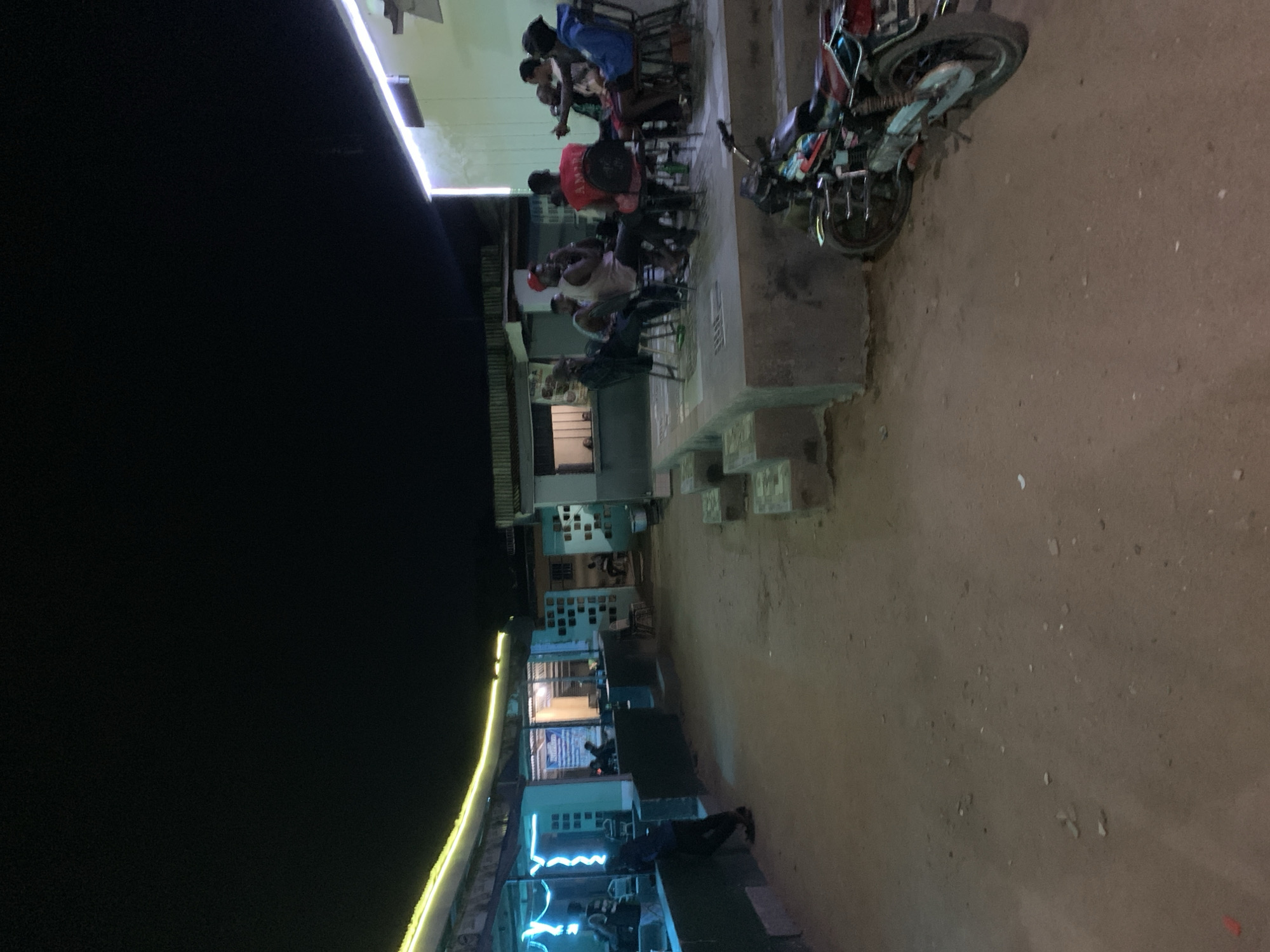 The image depicts a nighttime scene in New Stadium, Nigeria. Several people are sitting on chairs outside a building illuminated by LED strip lighting. A motorcycle is parked nearby. The ground is dirt. The atmosphere appears relaxed.