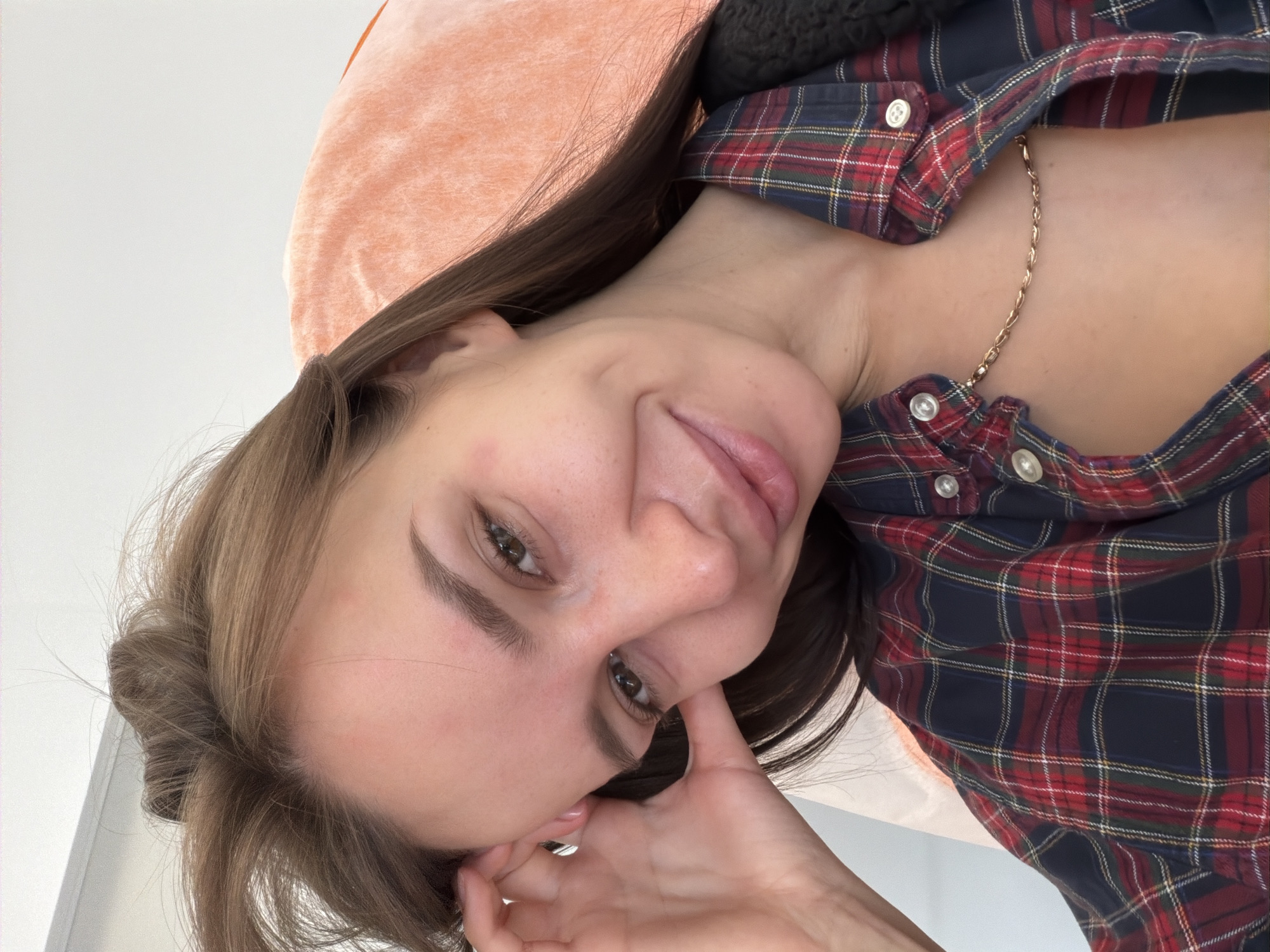 A person with fair skin, light brown hair, and brown eyes is looking at the camera with a slight smile. They are wearing a red, blue, and green plaid shirt with white buttons and a gold necklace. Their left hand is near their face. A peach-colored object is in the upper left corner. The location and city are not ascertainable from this image.