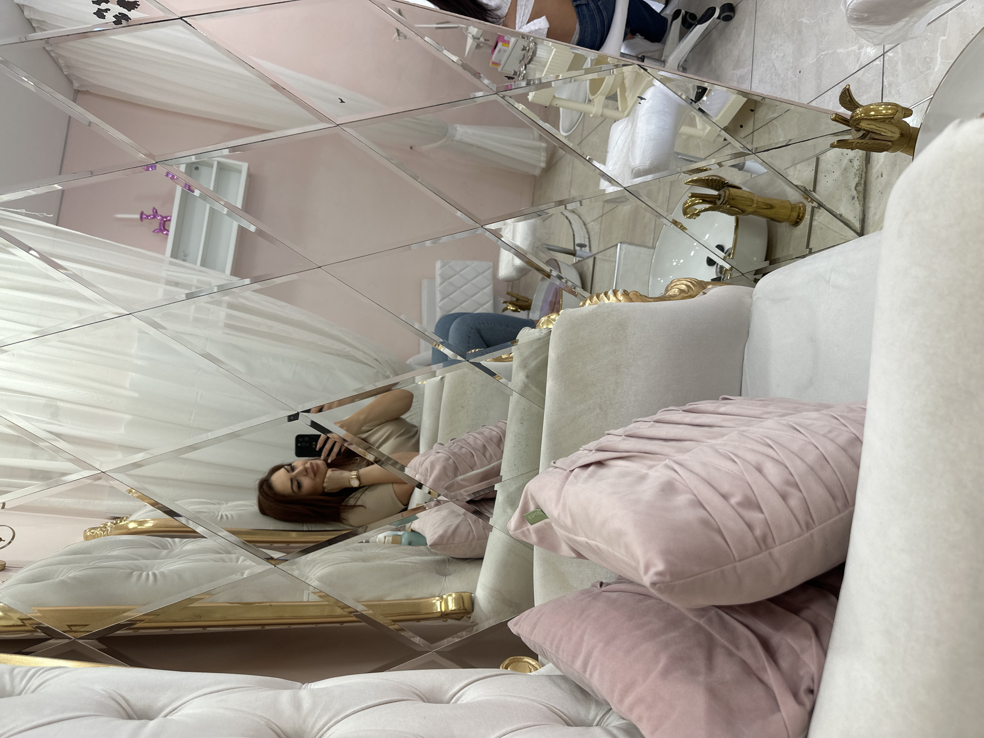 A woman is lying on a white tufted sofa and taking a photo of herself with her phone. She is in a room with a diamond-patterned mirrored wall, which reflects a pink-toned interior with a white sheer curtain, white shelving, and what appears to be manicure equipment. The room is decorated in a luxurious style with gold accents. The scene is in Sofia, Bulgaria.
