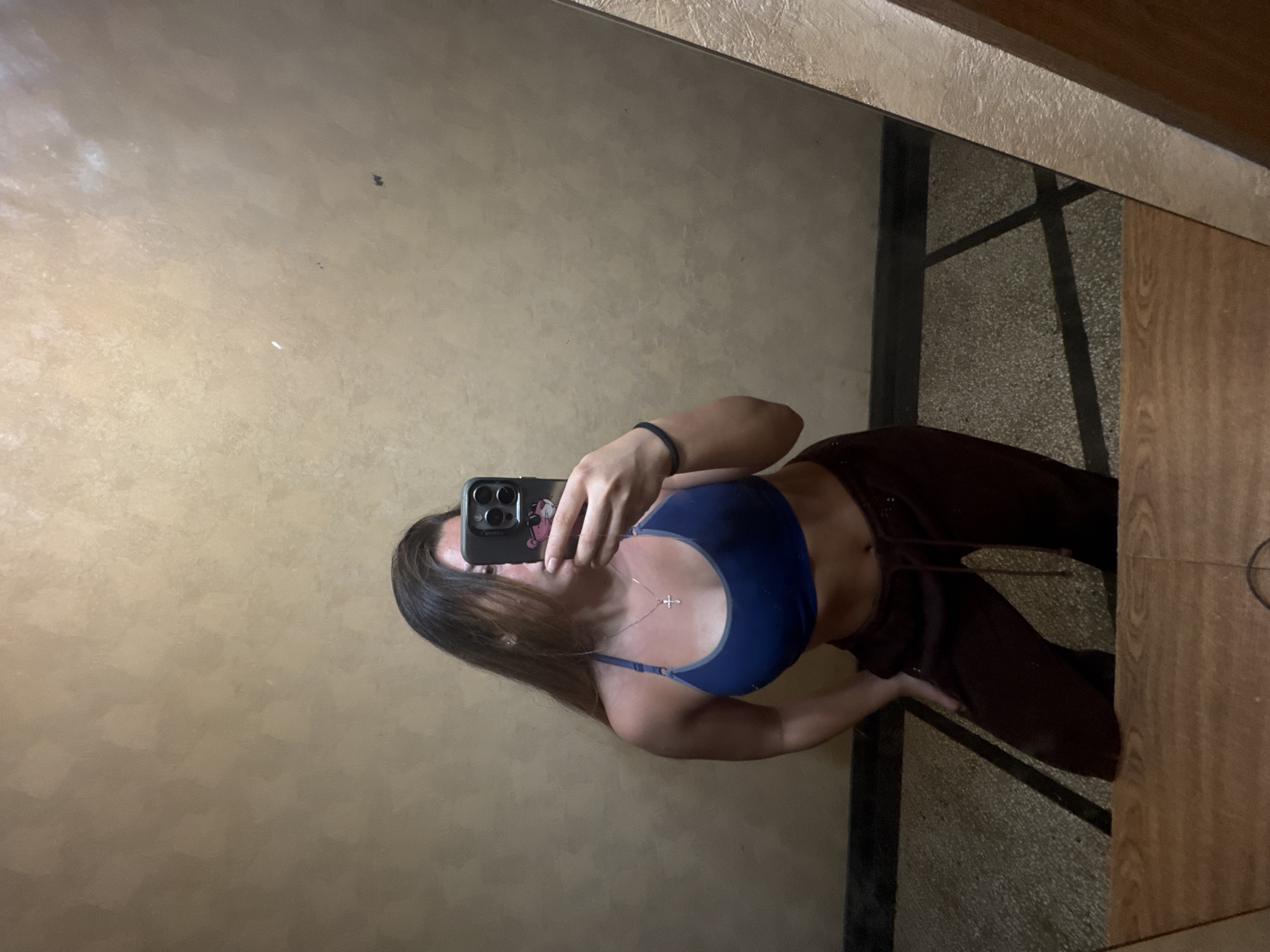 A young woman with long brown hair is taking a mirror selfie in what appears to be an indoor hallway. She is wearing a blue sports bra and dark maroon sweatpants. She has a silver necklace with a cross pendant and a black bracelet on her left wrist. Her right hand is resting on her right hip, and her left hand is holding her phone, which has a pink cartoon case on it. The hallway walls are a textured beige, and the floor is tiled with a darker pattern. There is a wooden railing in the foreground to the right. The scene does not provide any visual cues to suggest a location in Sofia, Bulgaria.