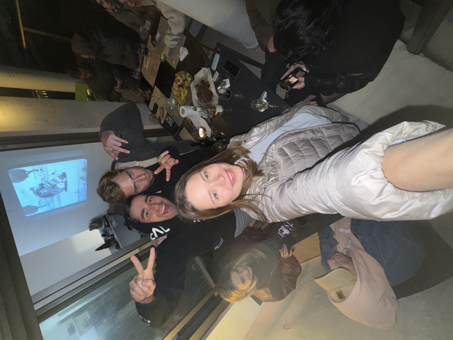 A group of people are crowded together, posing for a selfie. There are at least four people visible: three men and a woman holding the camera. One man is making a peace sign. A wall with a cartoon projection on it is visible to the left. There is a table with drinks and snacks in the background with more people milling around it. The image appears to be taken indoors at a party or social gathering.