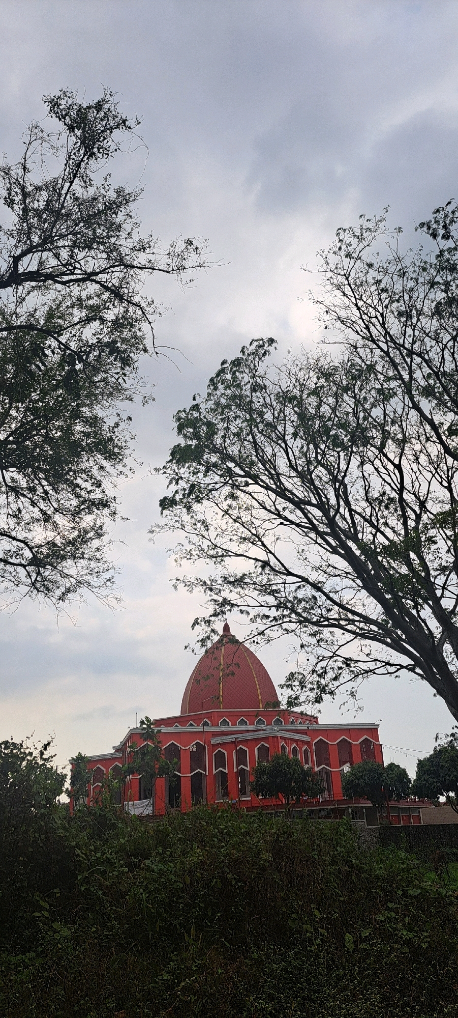 The image shows a red building with a dome, likely a mosque, located in Bulukandang, Indonesia. The building is partially obscured by the branches of trees in the foreground. The sky is overcast, suggesting a cloudy day. The building has a distinct architectural style with arched windows and decorative elements. Green foliage is visible in front of the building and surrounding it.