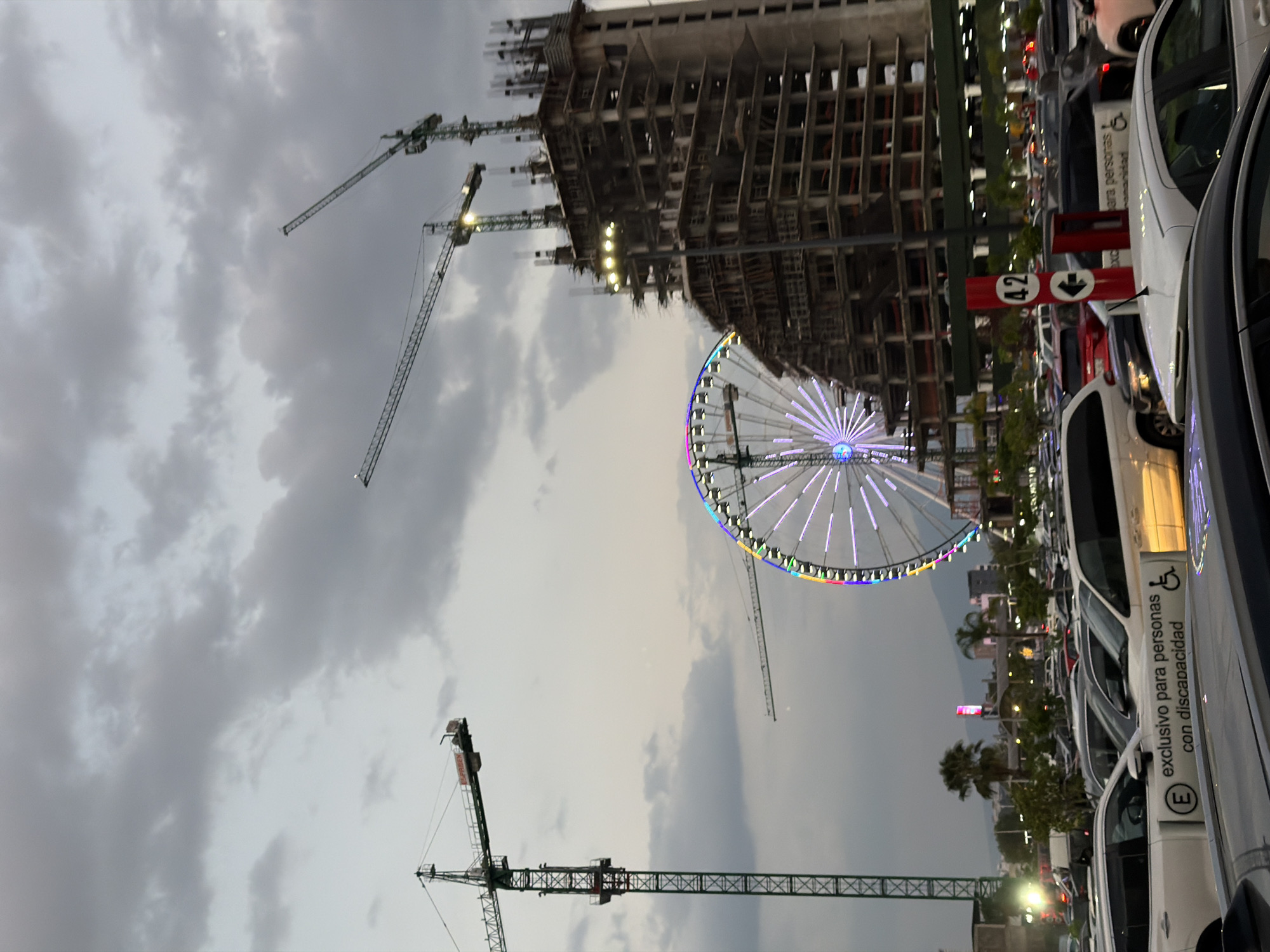 The image depicts an outdoor urban scene in Puebla, Mexico, showing a construction site with a building under construction, marked by multiple cranes. A Ferris wheel, lit with colorful lights, is positioned near the building. The foreground features a parking lot filled with vehicles, including cars marked for individuals with disabilities. The sky is overcast with gray clouds.