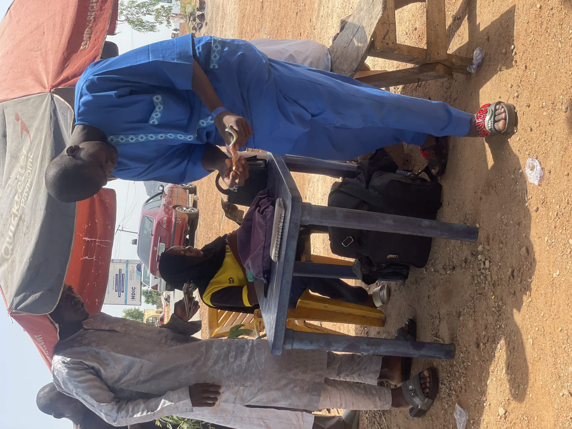 In Bauchi, Nigeria, under a red and white umbrella, two men and a woman are gathered around a blue table. One man, wearing a light-colored traditional garment, stands on the left, holding what appears to be a phone. Another man, dressed in a vibrant blue traditional outfit, stands on the right, examining money in his hands. The woman, seated, wears a yellow shirt with a black collar and a black headscarf. A yellow plastic chair supports her. On the table, a notebook, a purple pouch, and a black handbag are visible. Bags are also present on the ground. The scene unfolds on a dirt surface with scattered stones, under a clear, bright sky. A red car is parked in the background.